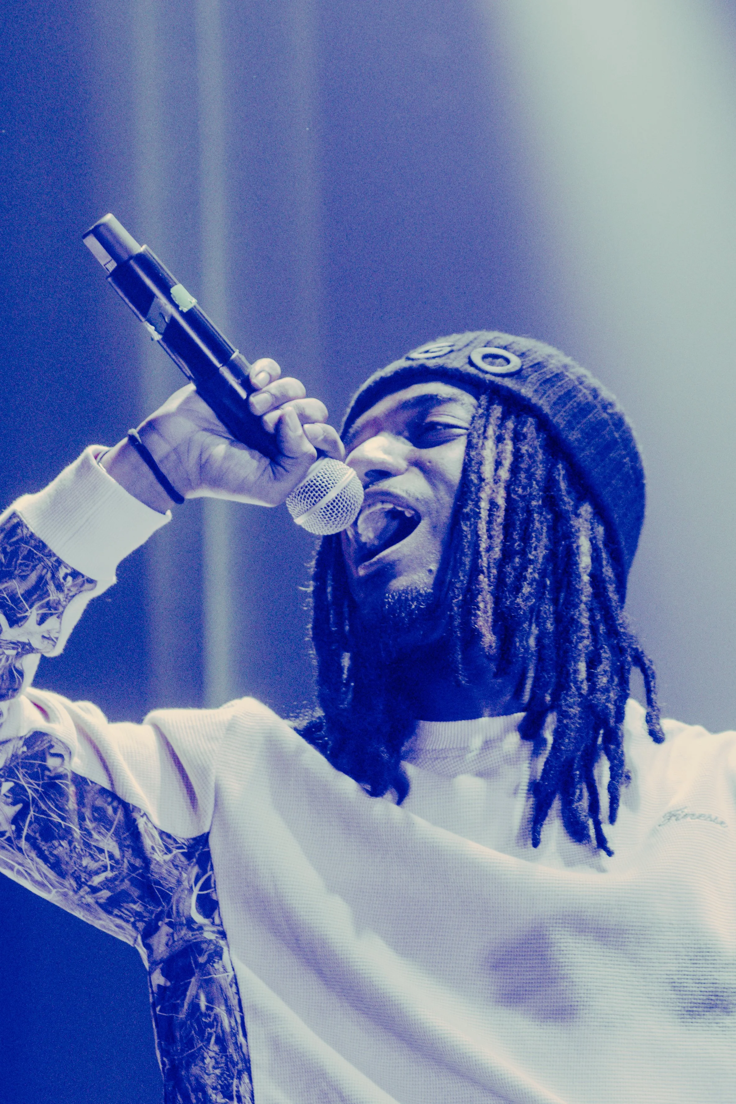Un homme avec des dreadlocks, portant un bonnet gris, chante dans un microphone sur scène, avec un éclairage bleuâtre.