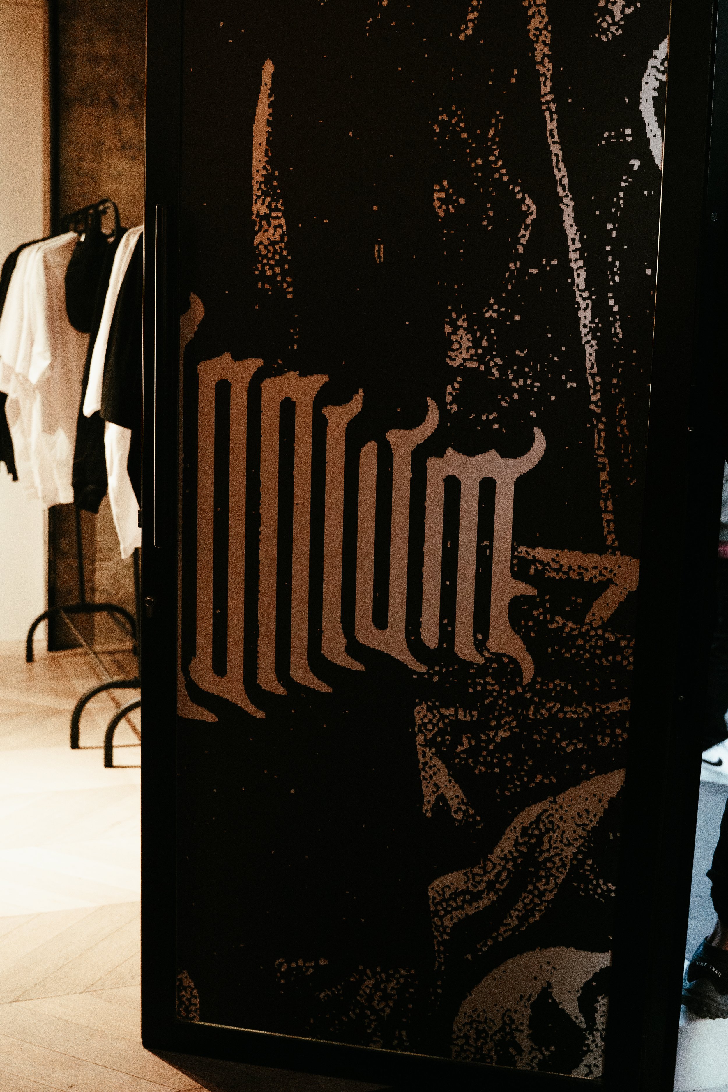 Cabinet noir avec un motif abstrait en teintes beige et brun, dans un intérieur avec des vêtements suspendus en arrière-plan.