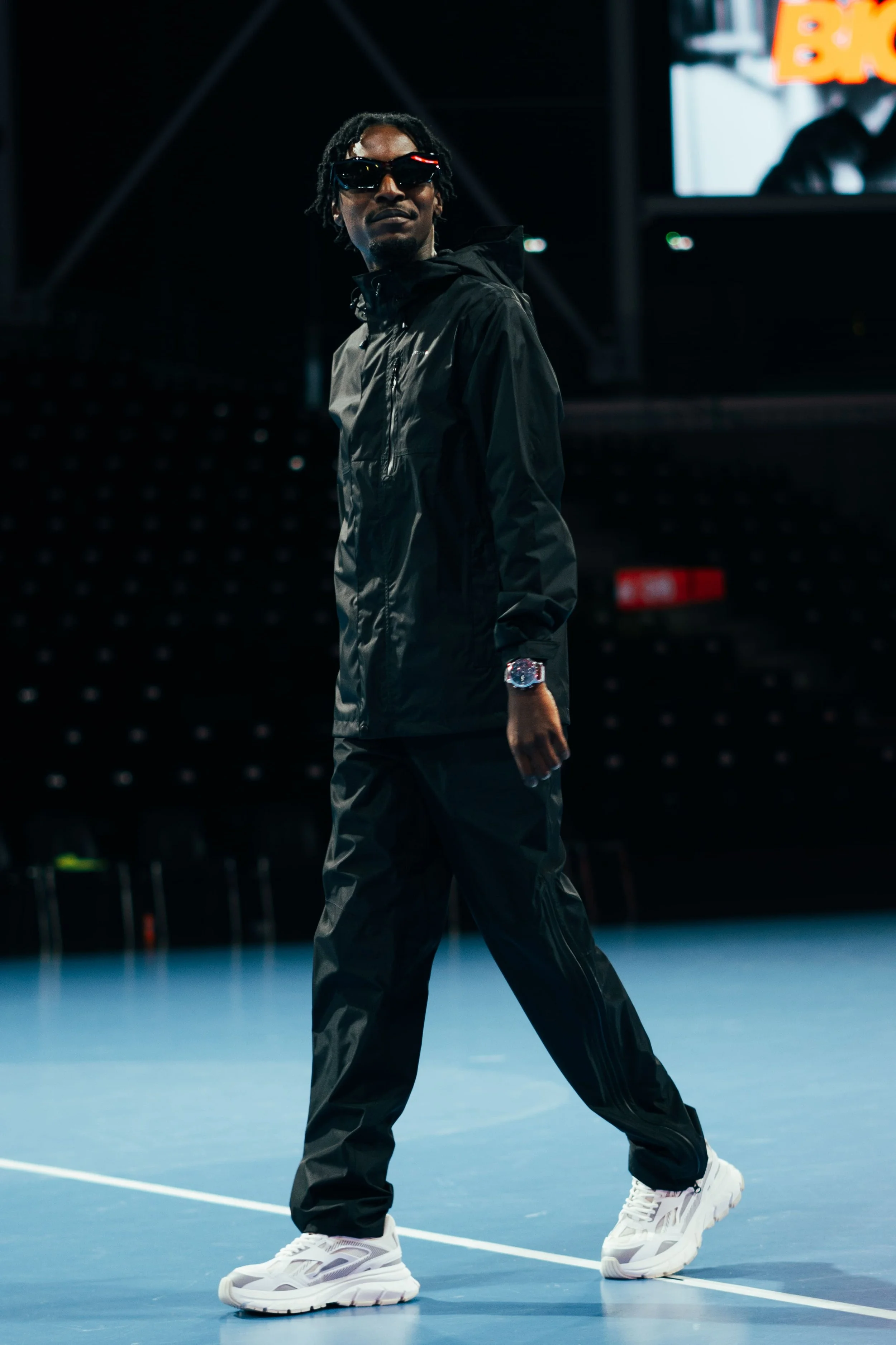 Homme en vêtements de sport noirs avec des baskets blanches sur un terrain de tennis intérieur, portant des lunettes de soleil.
