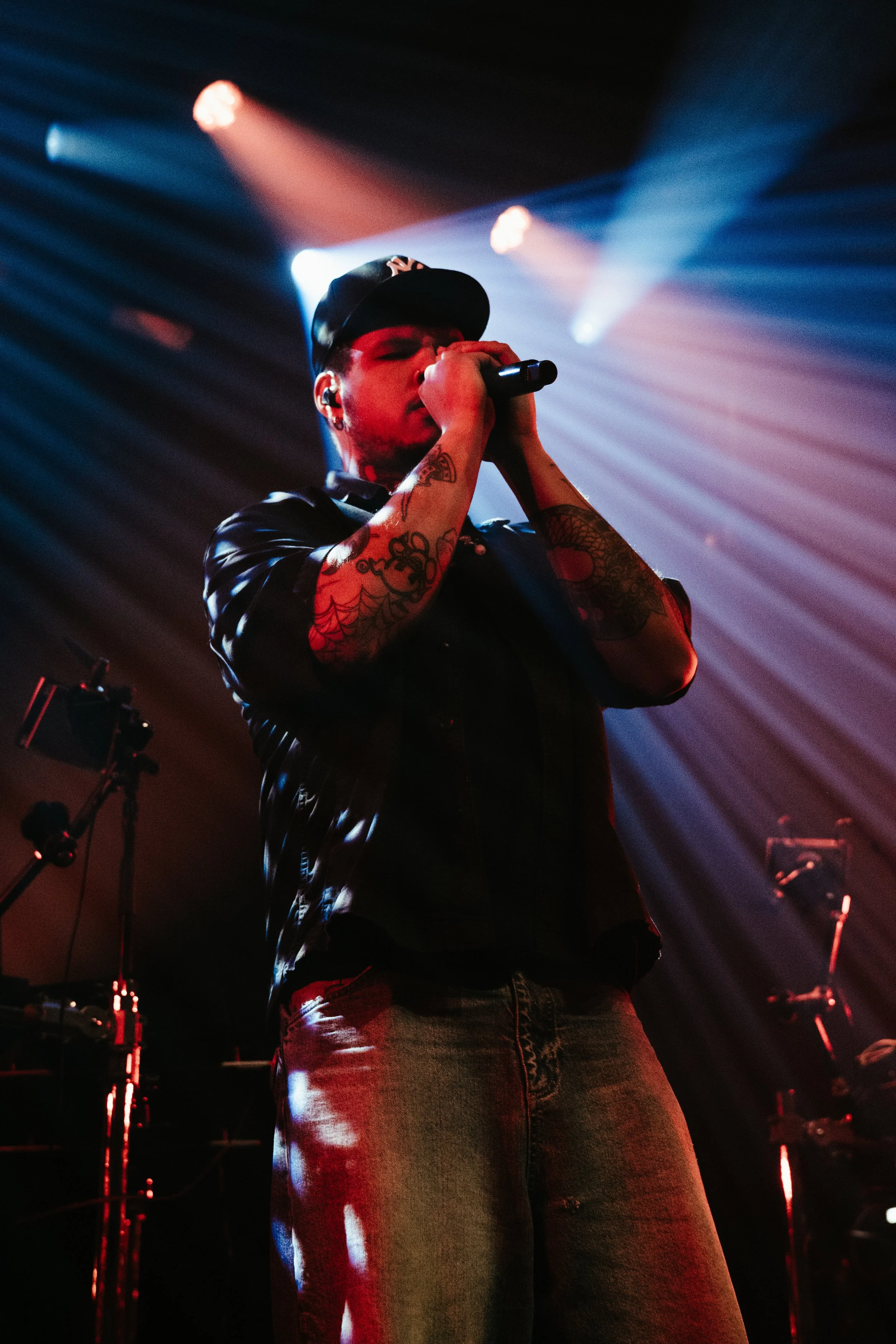Un chanteur portant une casquette noire et une veste en cuir chante dans un concert, avec un éclairage coloré en arrière-plan.