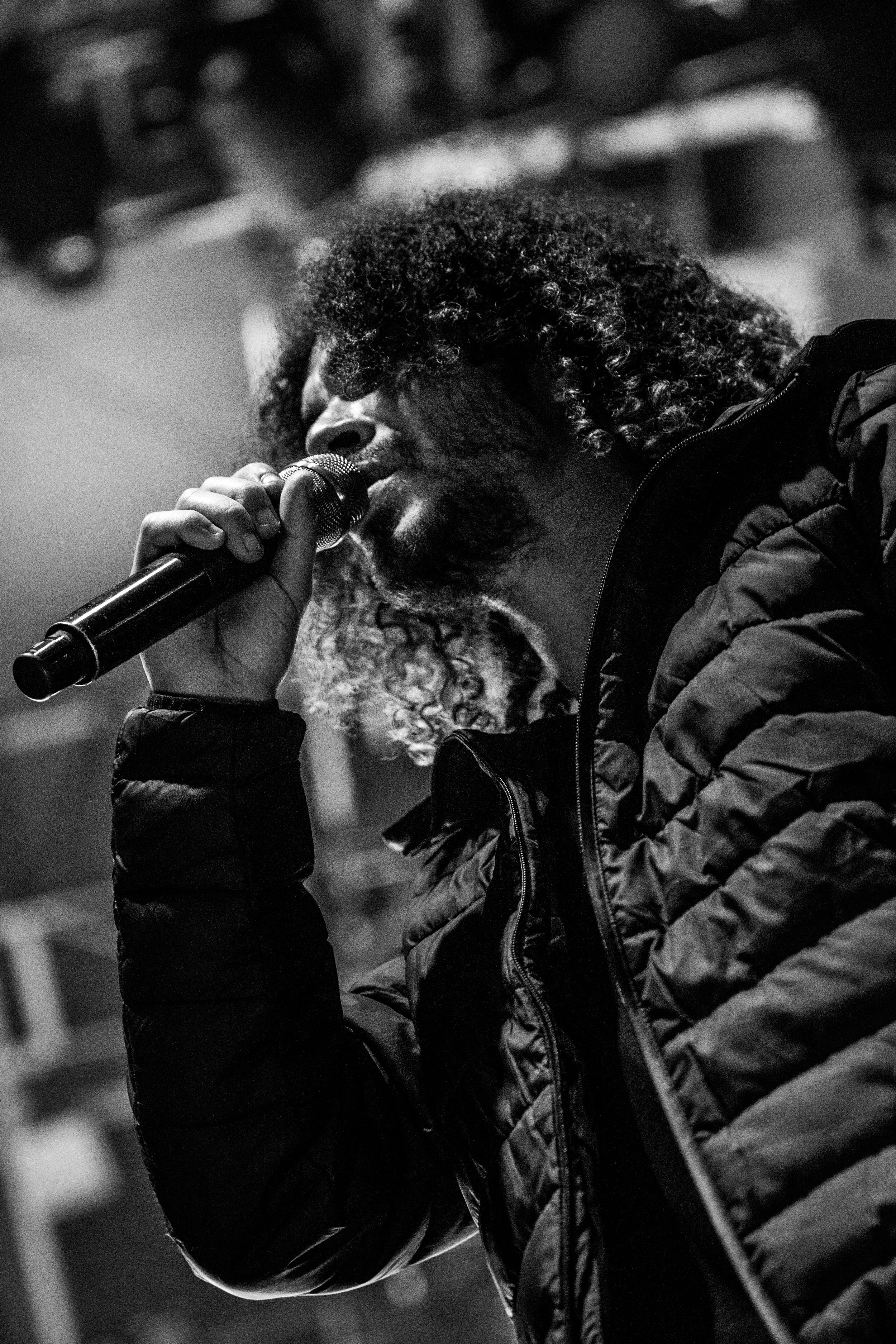 Un homme avec des cheveux bouclés tient un microphone et chante ou parle, vêtu d'une veste noire, dans une ambiance sombre.