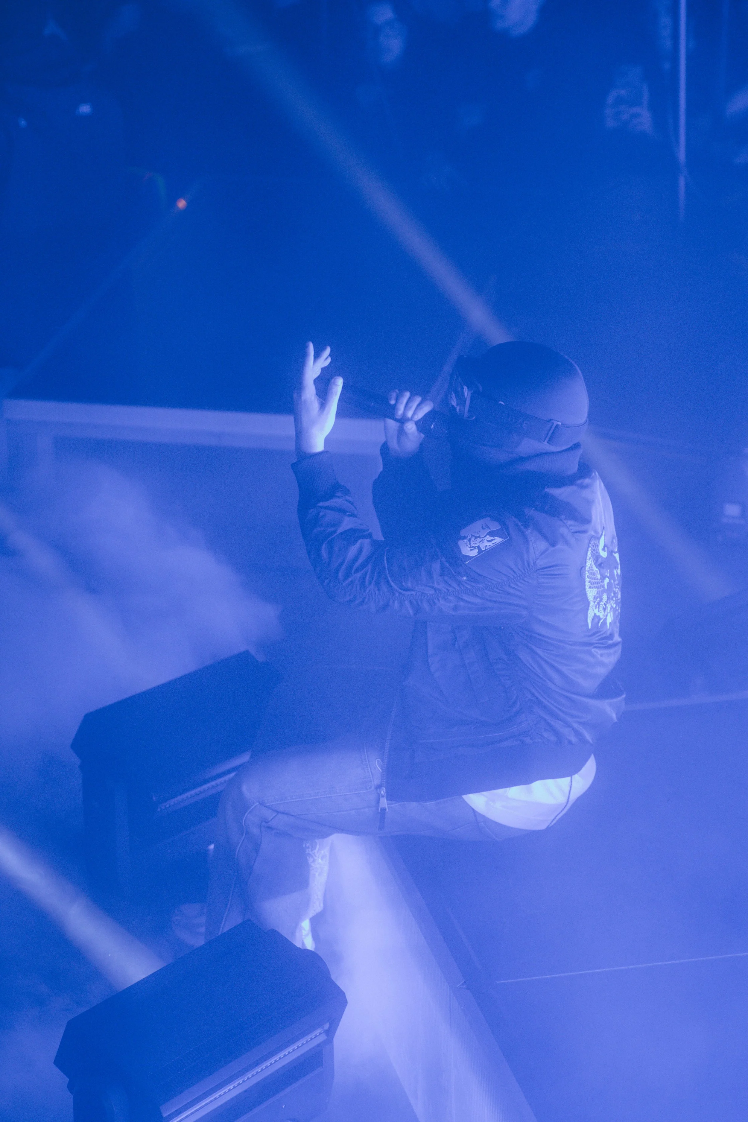Un artiste en costume noir joue du violon sur scène sous des lumières bleues et vaporeuses, vu de dos.