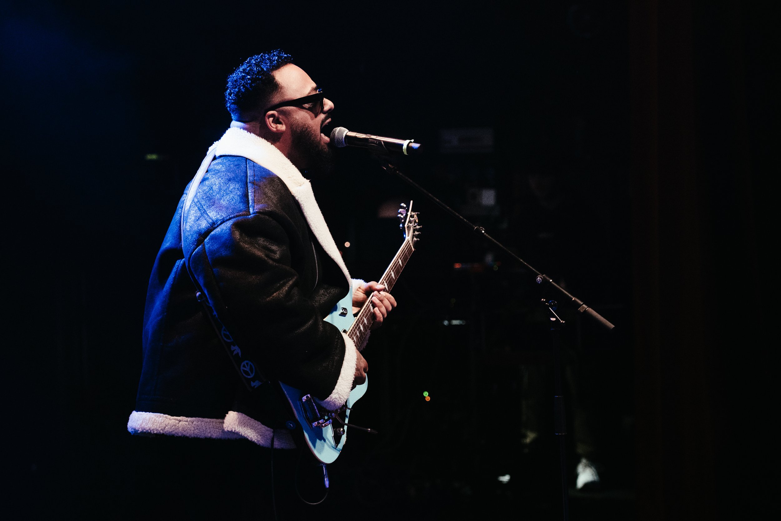 Un homme chante et joue de la guitare électrique sur scène, portant une veste en cuir avec pellicule blanche, en environnement sombre.