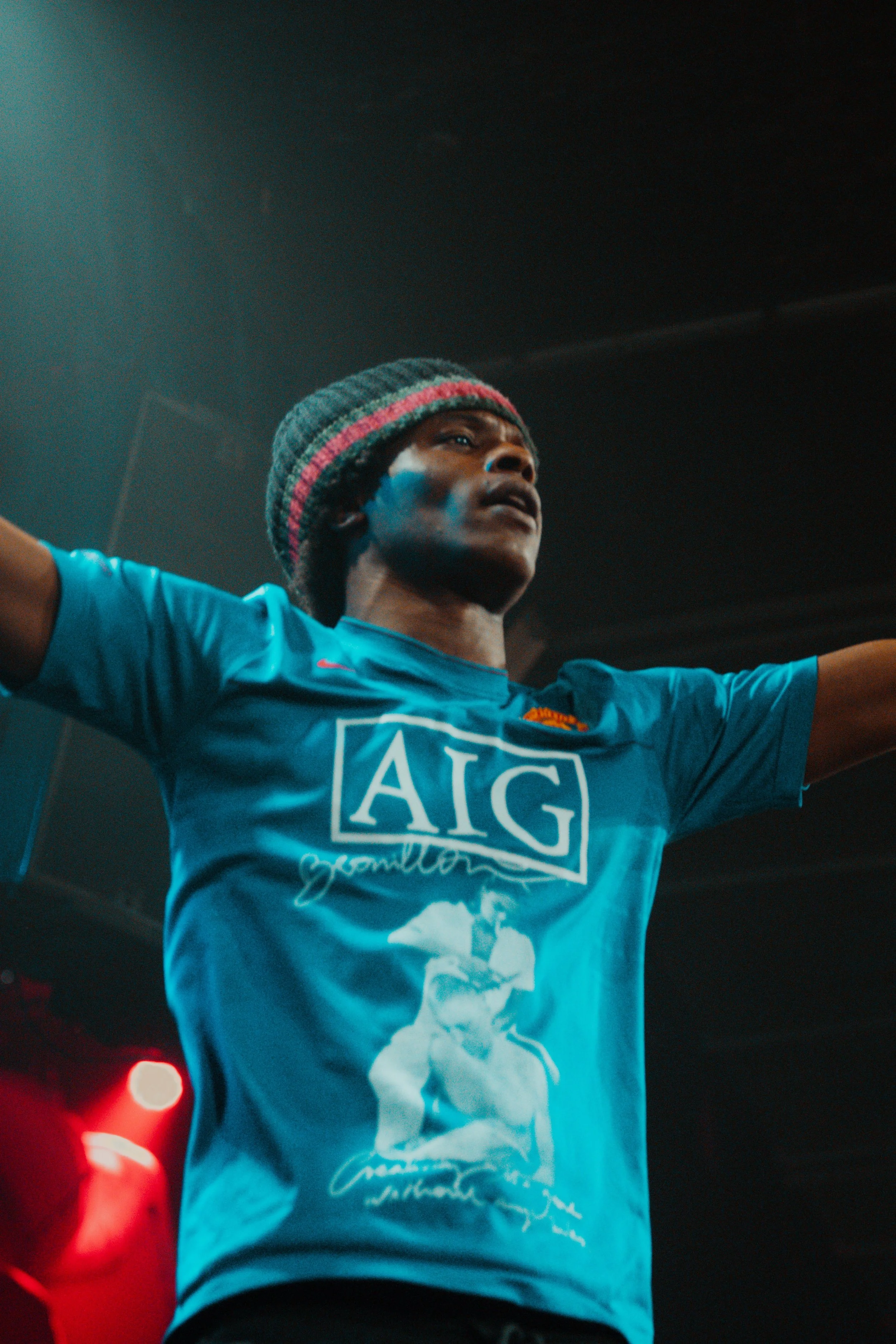 Jeune homme portant un maillot de sport bleu avec le logo AIG et une casquette en laine, levant les bras dans une salle sombre avec un éclairage rougeâtre.