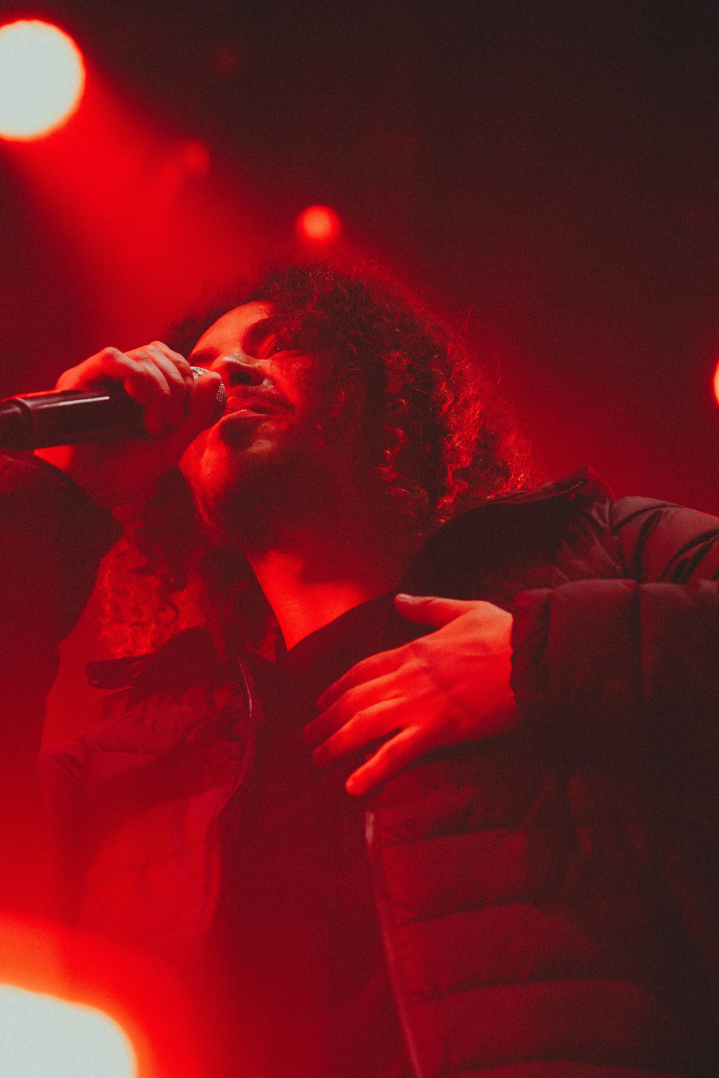 Un chanteur ou chanteuse avec des cheveux bouclés, tenant un microphone, sous un éclairage rouge sur scène.