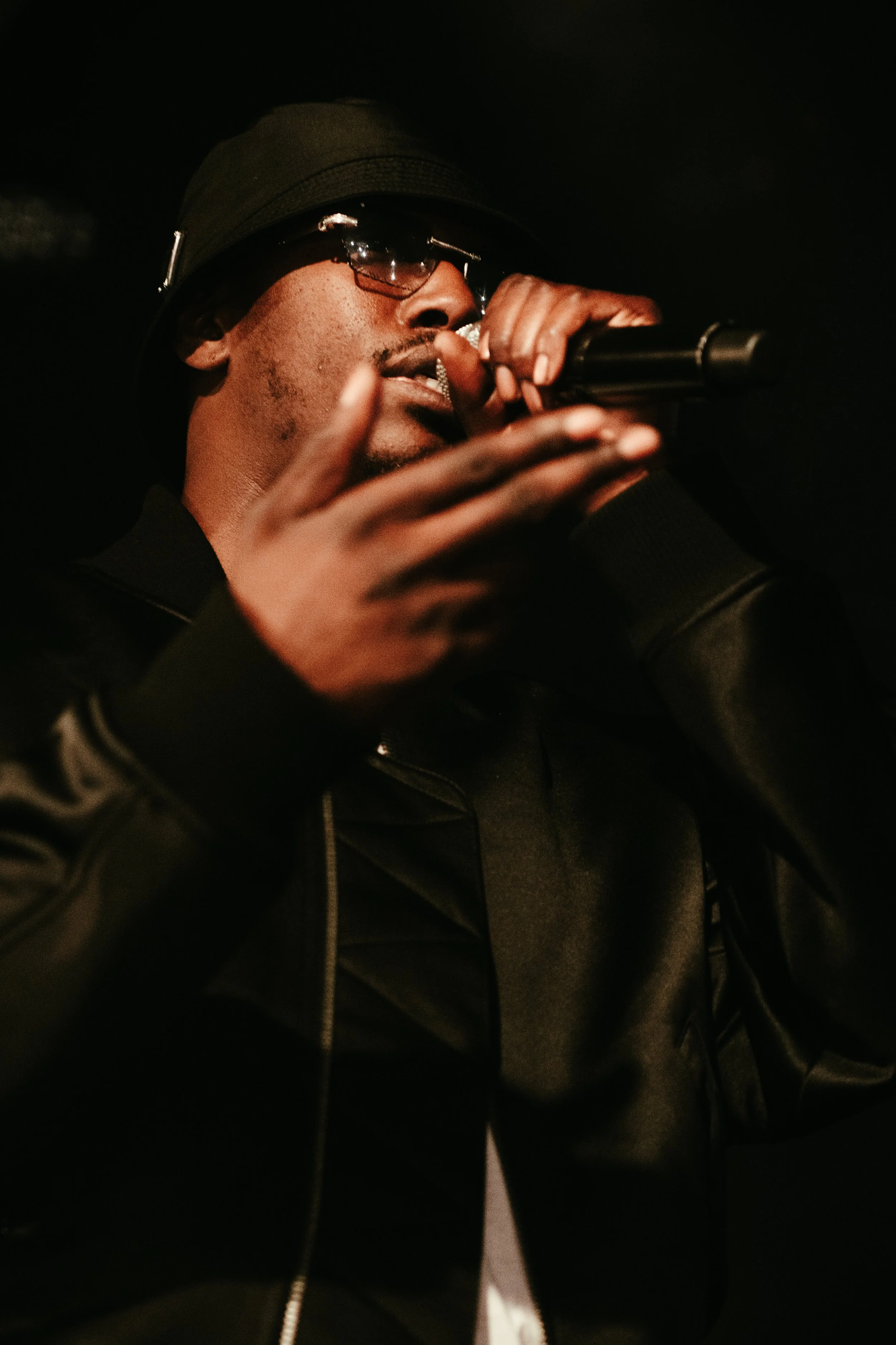 Un homme portant un chapeau noir et des lunettes de soleil chante ou parle dans un microphone, vêtu d'une veste en cuir noir, dans un environnement sombre.