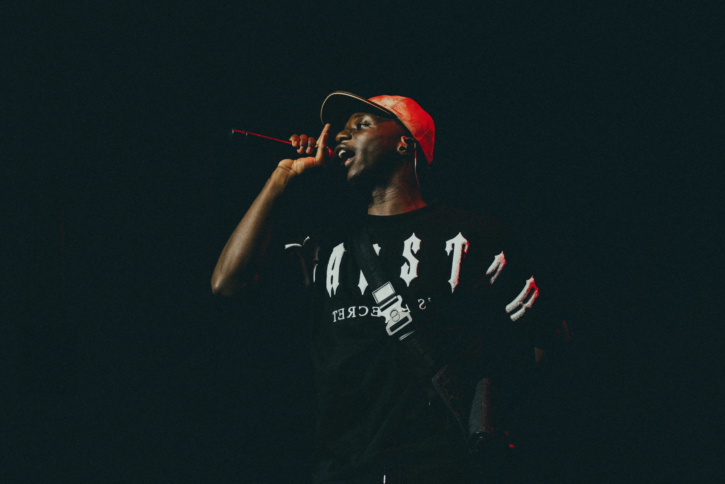 Un homme portant une casquette rouge, tenant un microphone, en train de chanter ou de parler, avec un fond sombre.