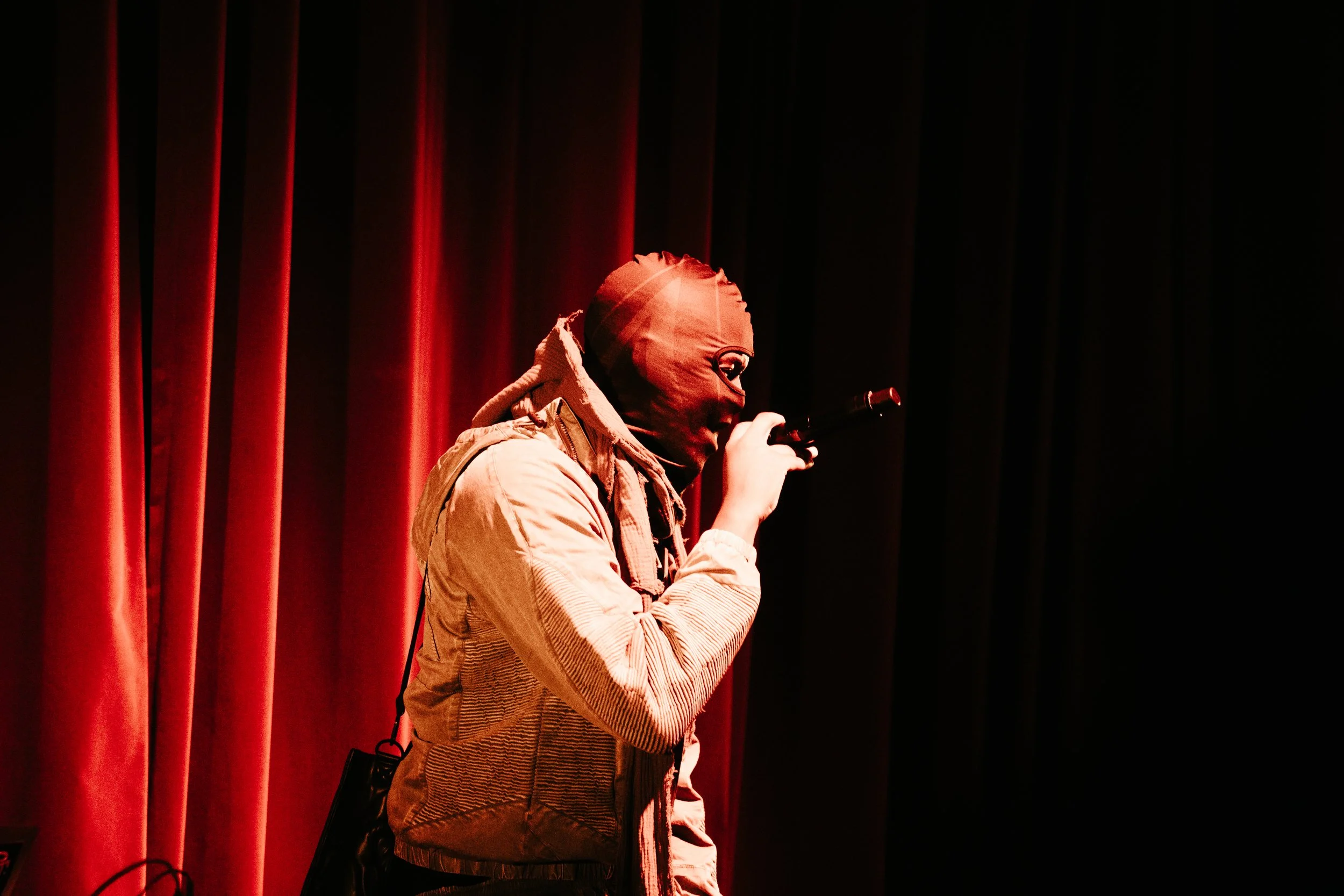 Personne portant un masque capsule noir, tenant un microphone, sur scène avec rideaux rouges.
