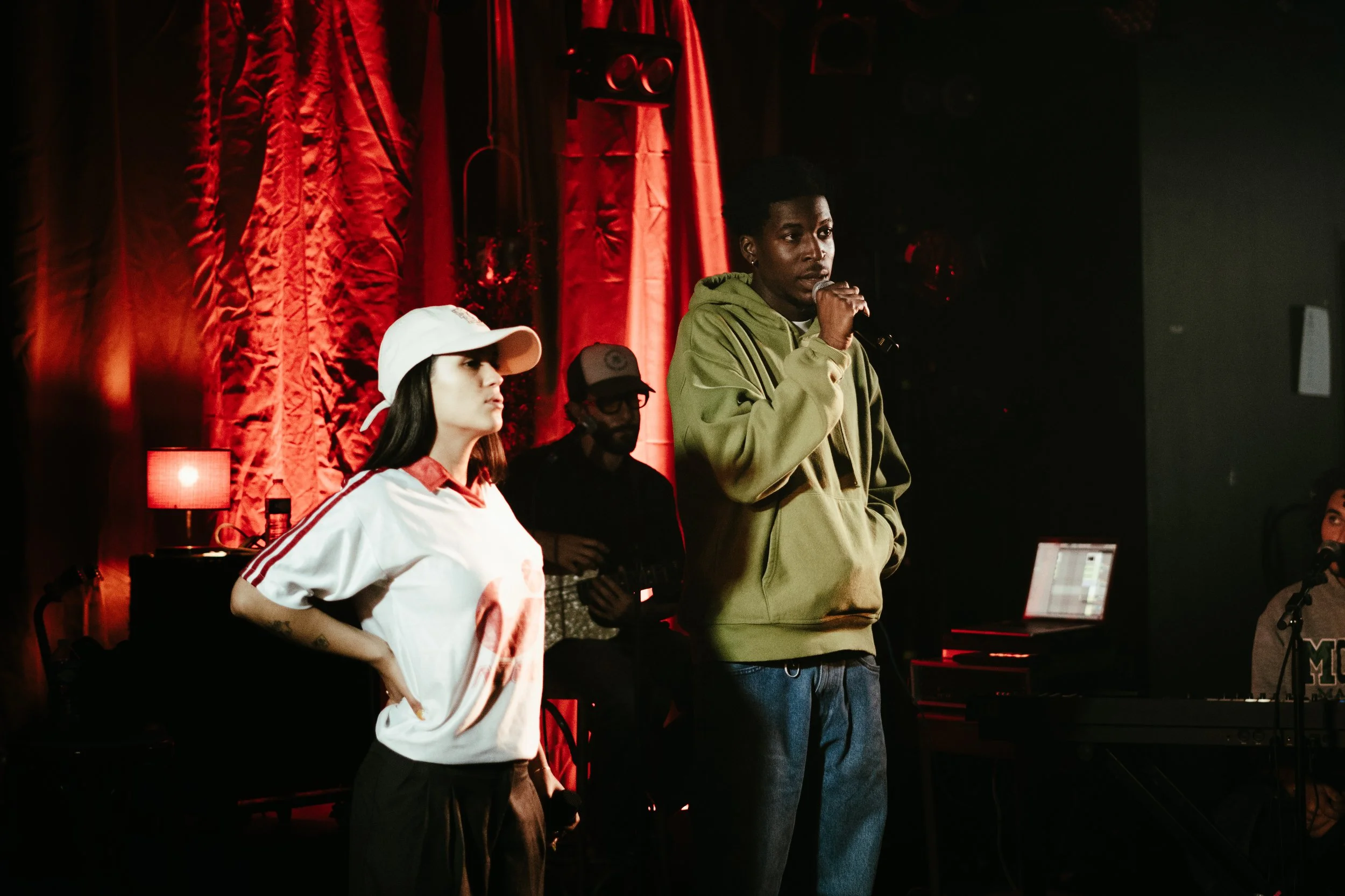 Un groupe de personnes sur scène lors d'une performance, avec un homme qui parle dans un microphone, entouré de plusieurs musiciens et un fond de rideaux rouges.