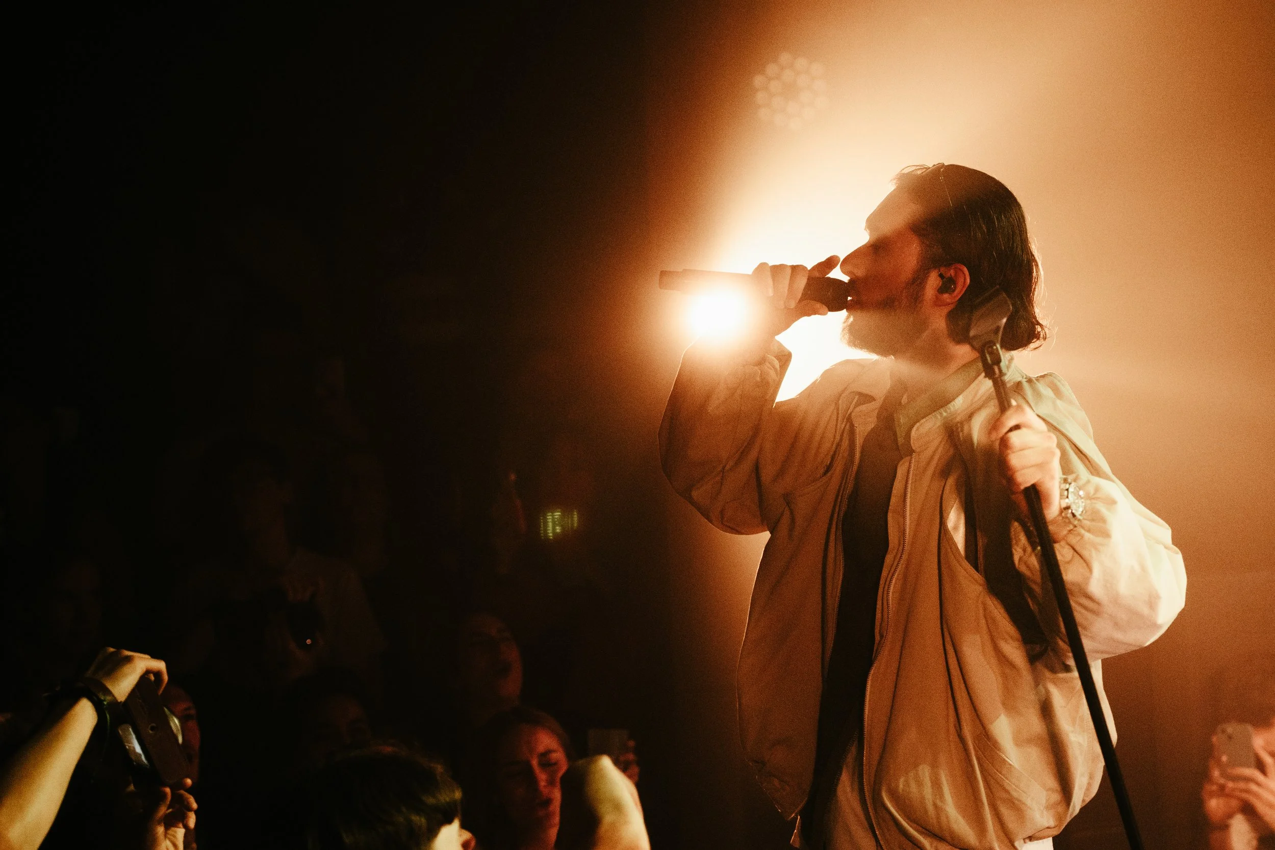 Un chanteur masculine avec une barbe et cheveux longs chantant dans un micro sur scène, éclairé par une lumière chaude, avec un public devant lui.