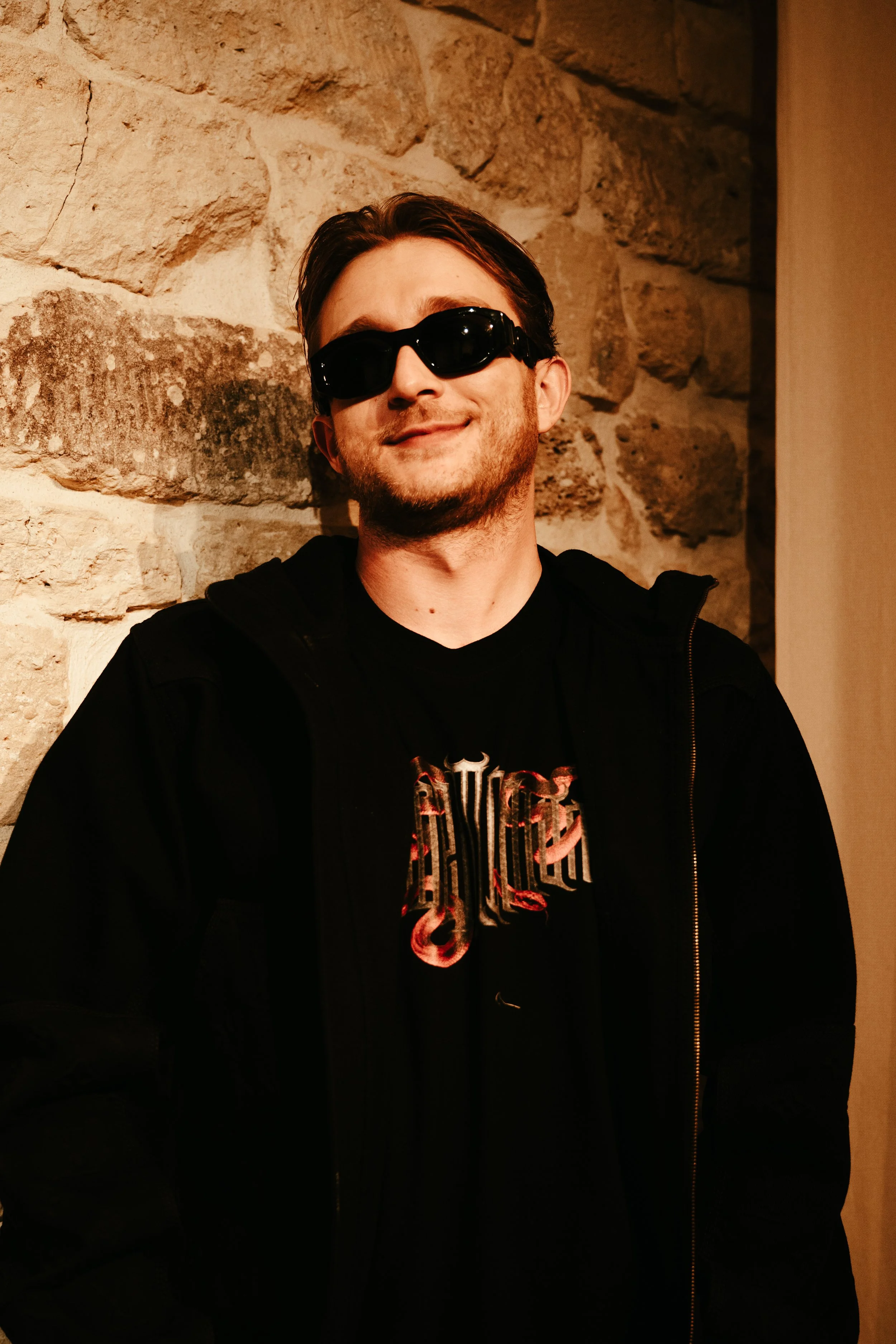 Homme souriant portant des lunettes de soleil noires, vêtement noir avec un t-shirt imprimé, devant un mur en pierre.