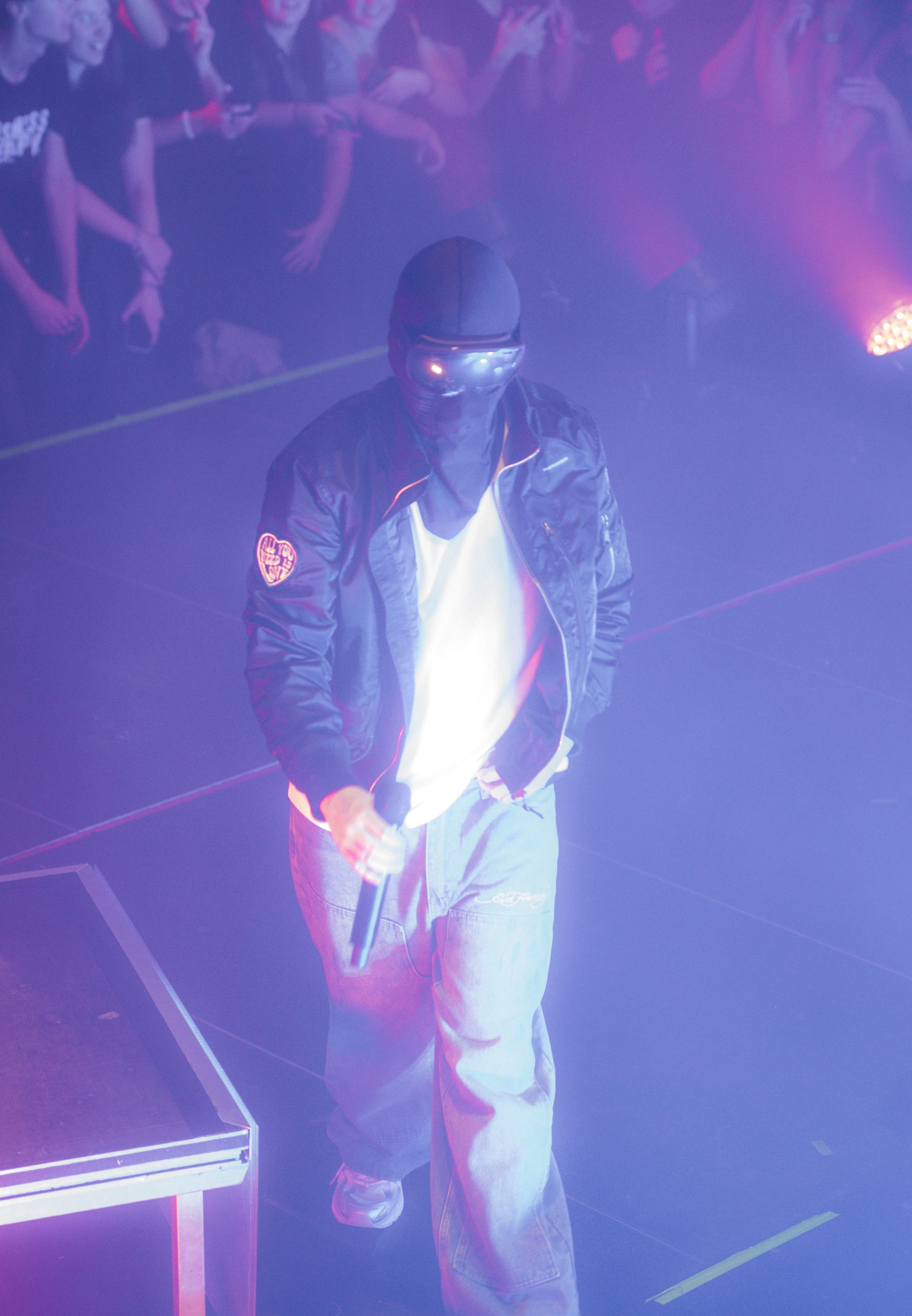 Un artiste portant un casque, une veste en cuir noire et un pantalon ample, tient un microphone lors d'une performance sur scène avec des lumières colorées et un public en arrière-plan.