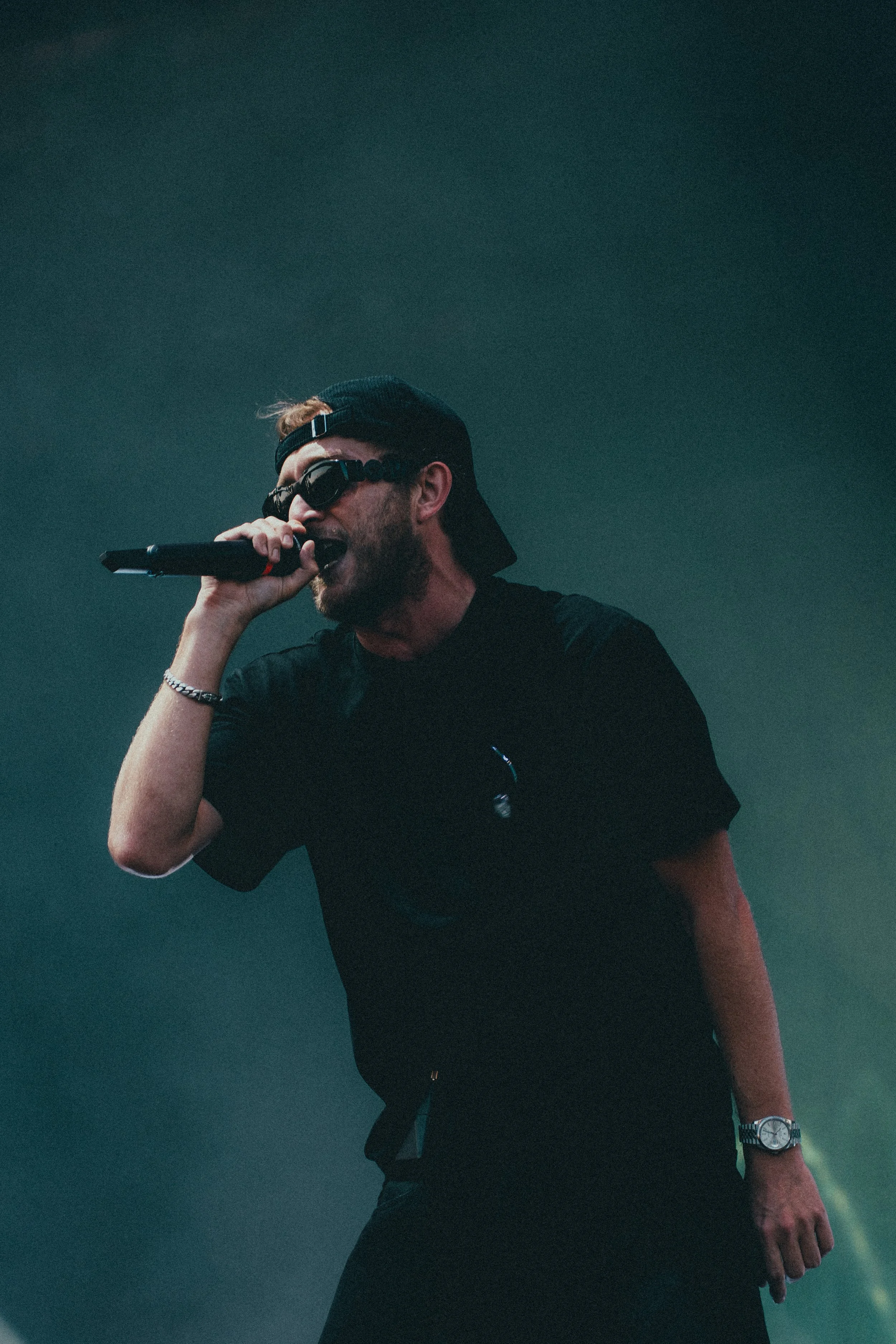 Un homme portant une casquette, des lunettes de soleil et un bracelet, tenant un microphone en pleine performance.