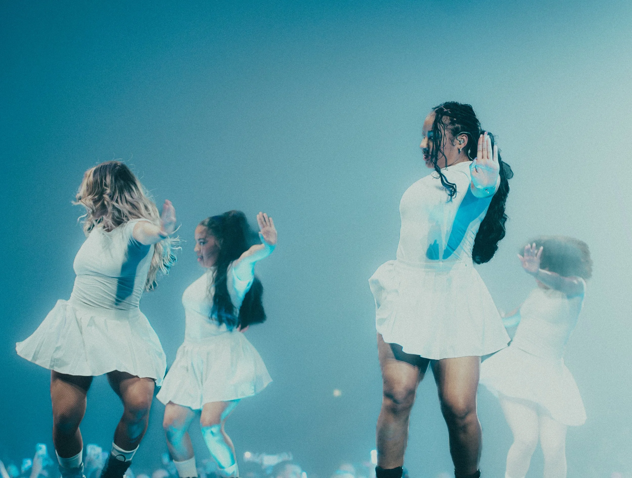 Quatre danseuses en blanc exécutant une chorégraphie sur scène, sous un éclairage bleuâtre, avec des spectateurs en arrière-plan.