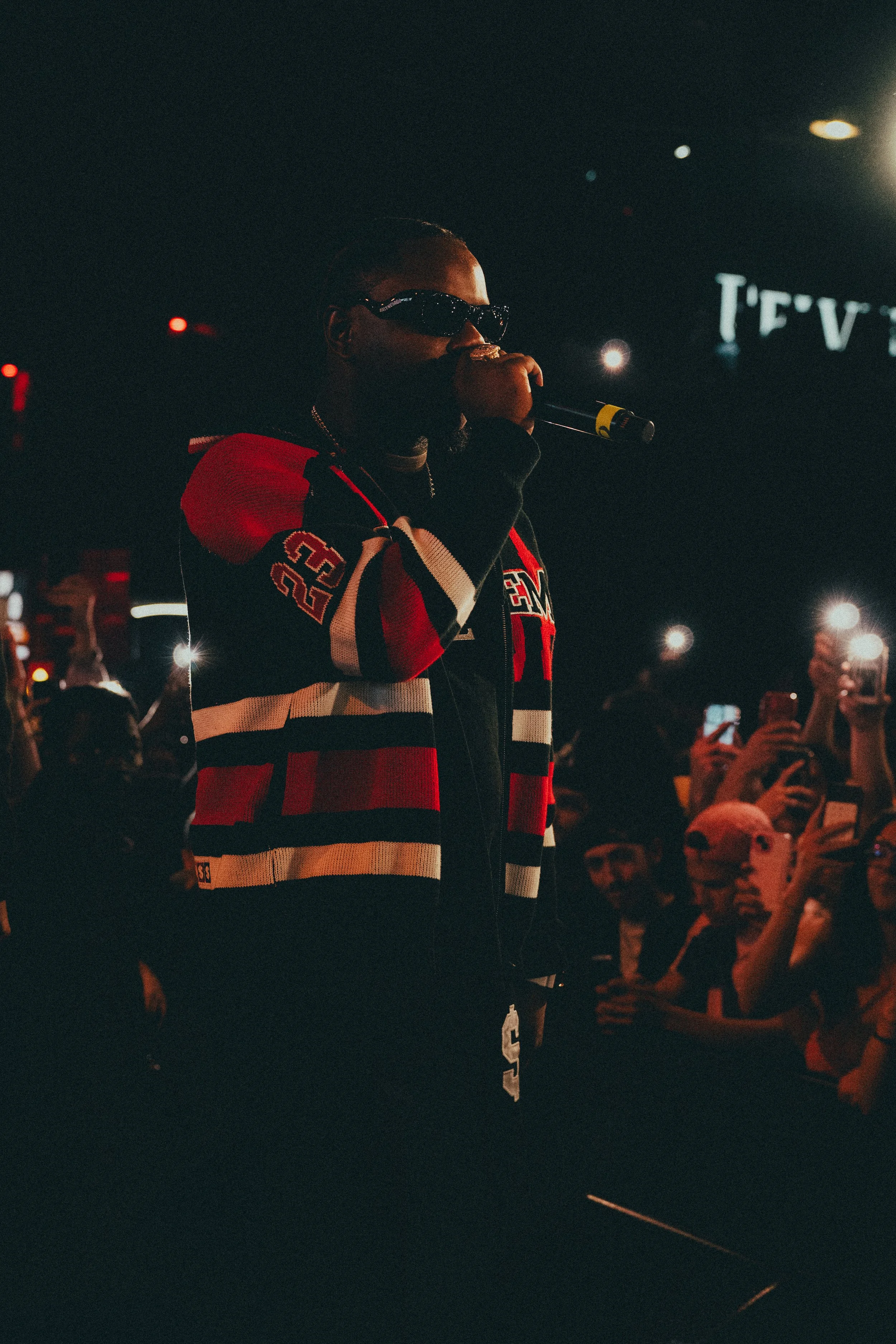 Un artiste en avant-plan portant une veste à rayures rouges, blanches et noires, et portant des lunettes noires, en train de chanter ou parler dans un micro. En arrière-plan, une foule de personnes avec des téléphones, dans une ambiance sombre avec d