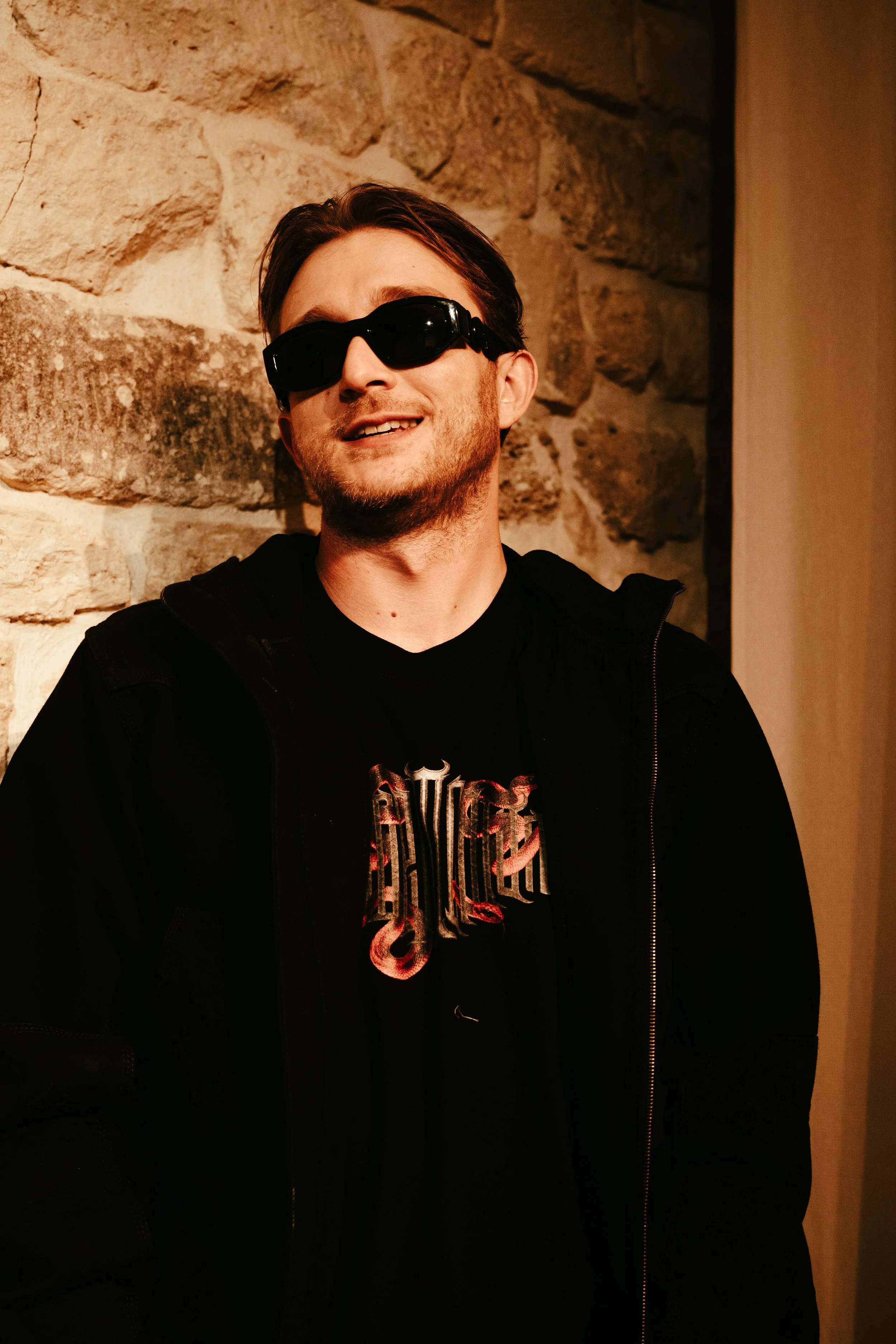 Jeune homme portant des lunettes de soleil noires, vêtu d'un t-shirt noir avec un logo métallique et d'une veste noire, posant devant un mur en pierre.