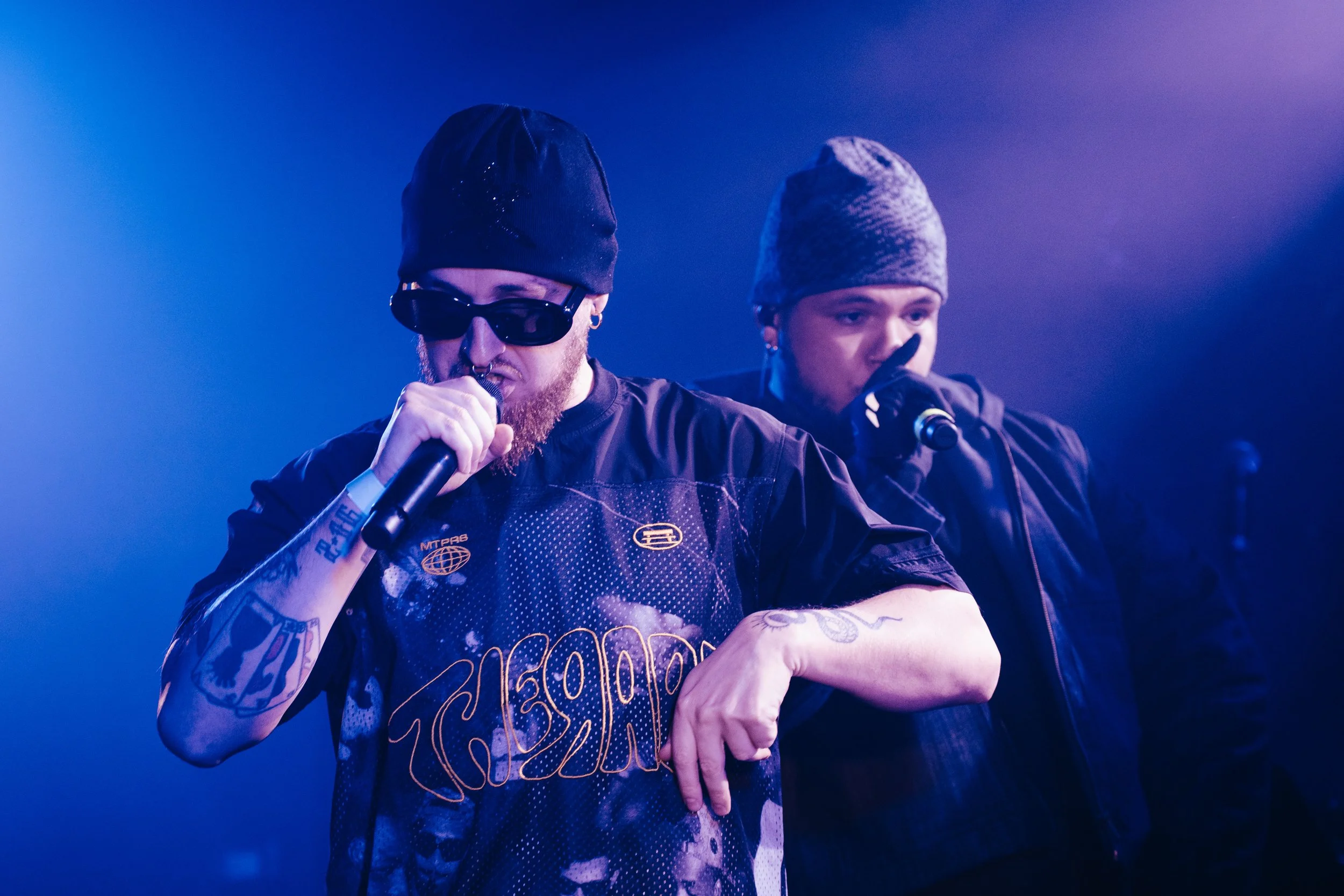 Deux rappeurs sur scène, portant des bonnets, en train de chanter dans un micro, avec des lumières violettes et bleues.