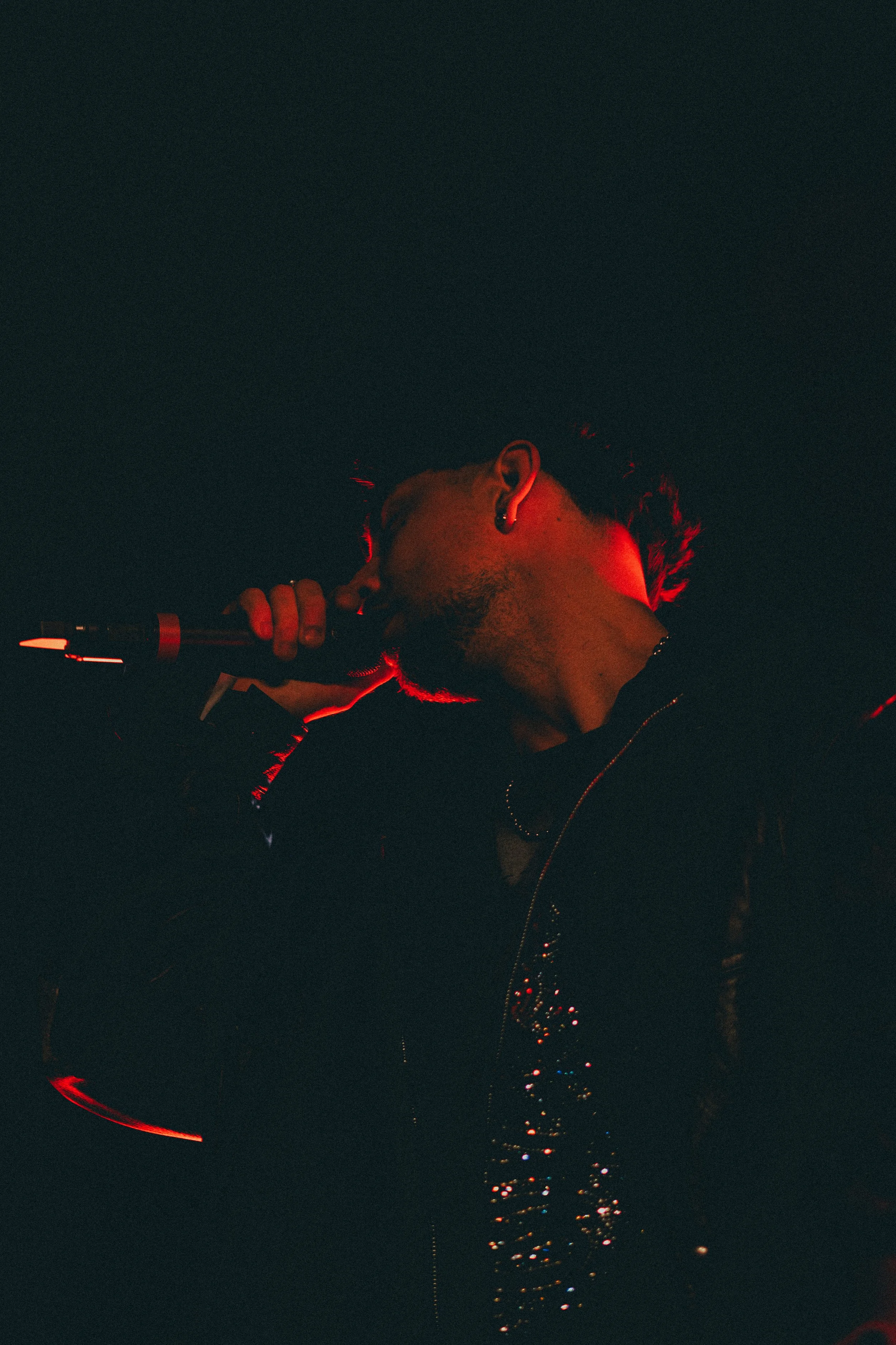 Un homme chante avec un micro dans une ambiance sombre et rouge, portant des écouteurs et un vêtement noir.