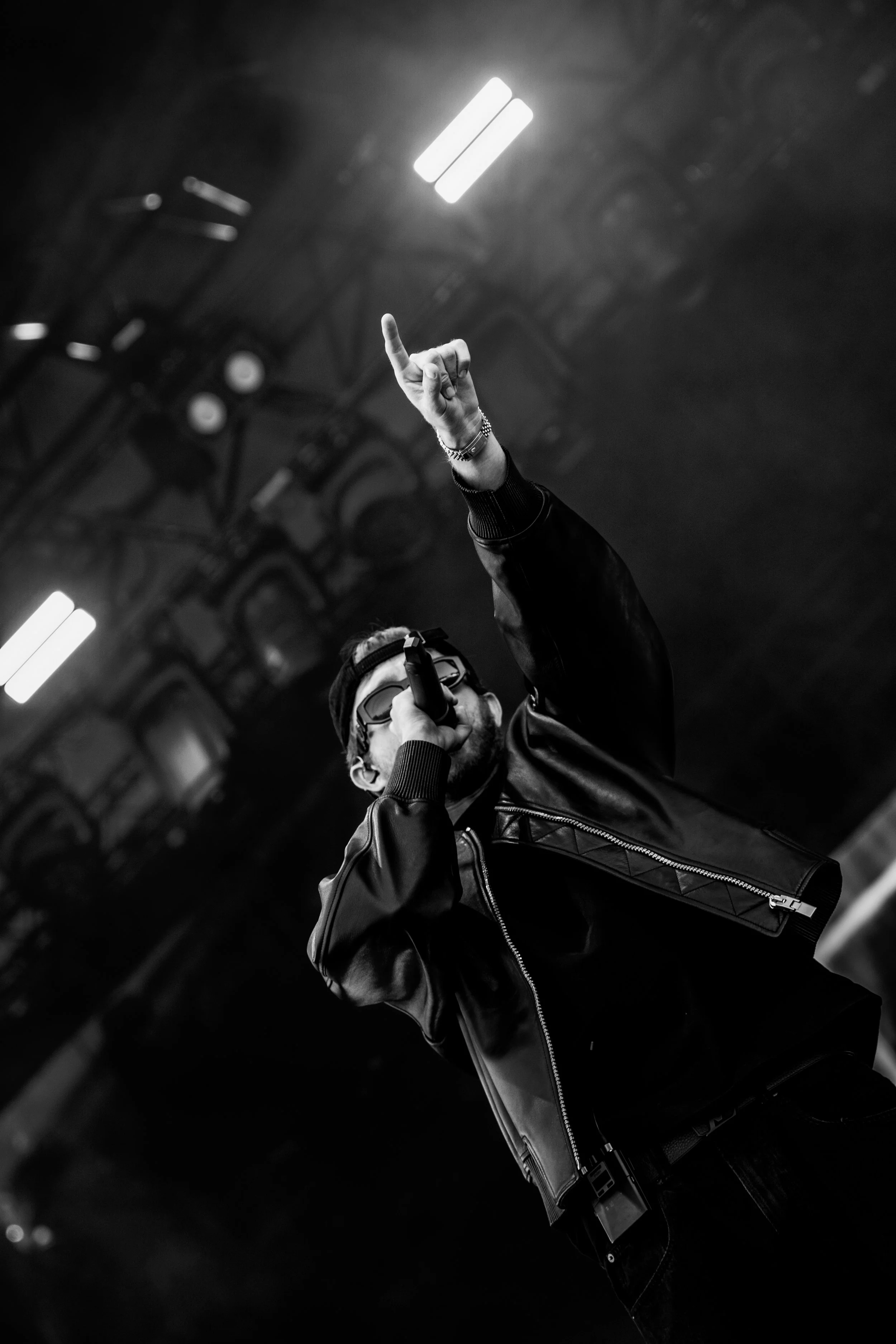 Un chanteur masculi sol en noir et blanc, portant des lunettes de soleil, une veste en cuir, tenant un microphone, et levant le doigt en l'air lors d'une performance sur scène