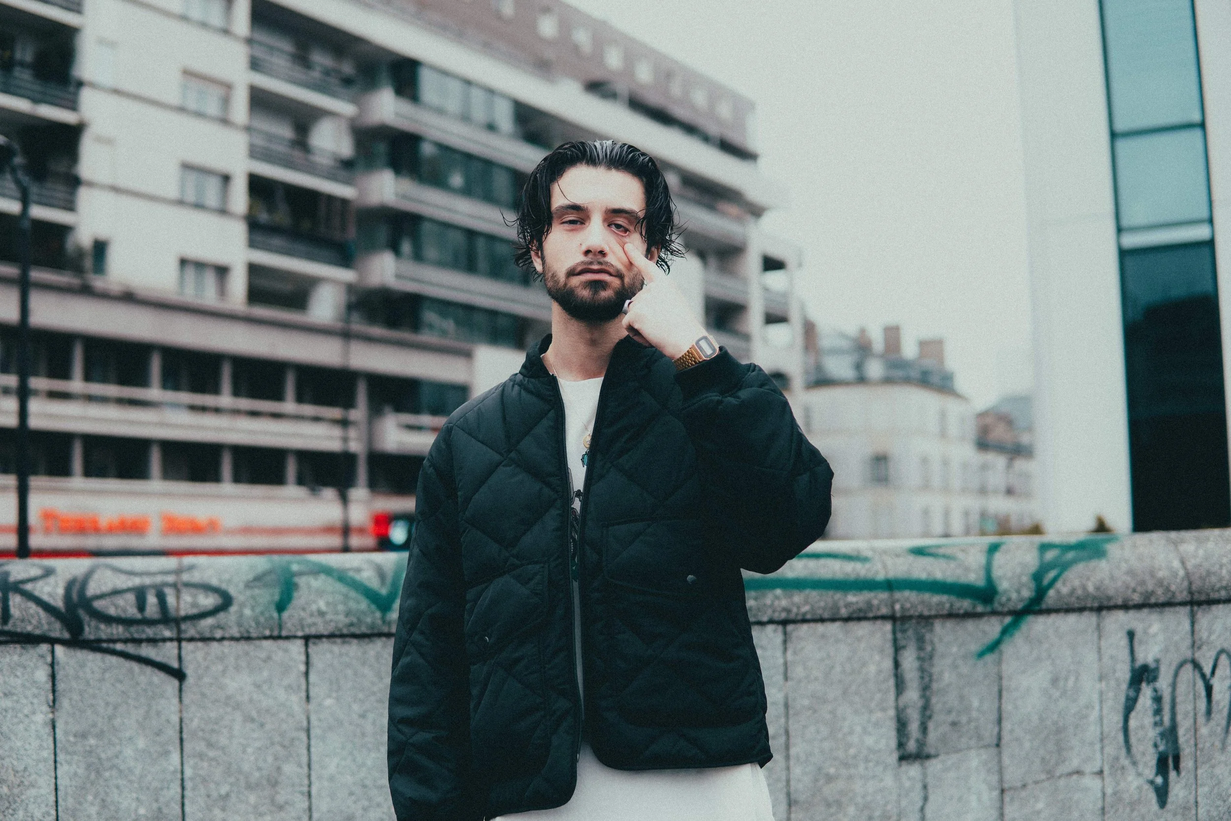 Un homme avec les cheveux noirs, barbe, portant une veste noire et un smartwatch, se tient devant un mur graffitié dans une ville.