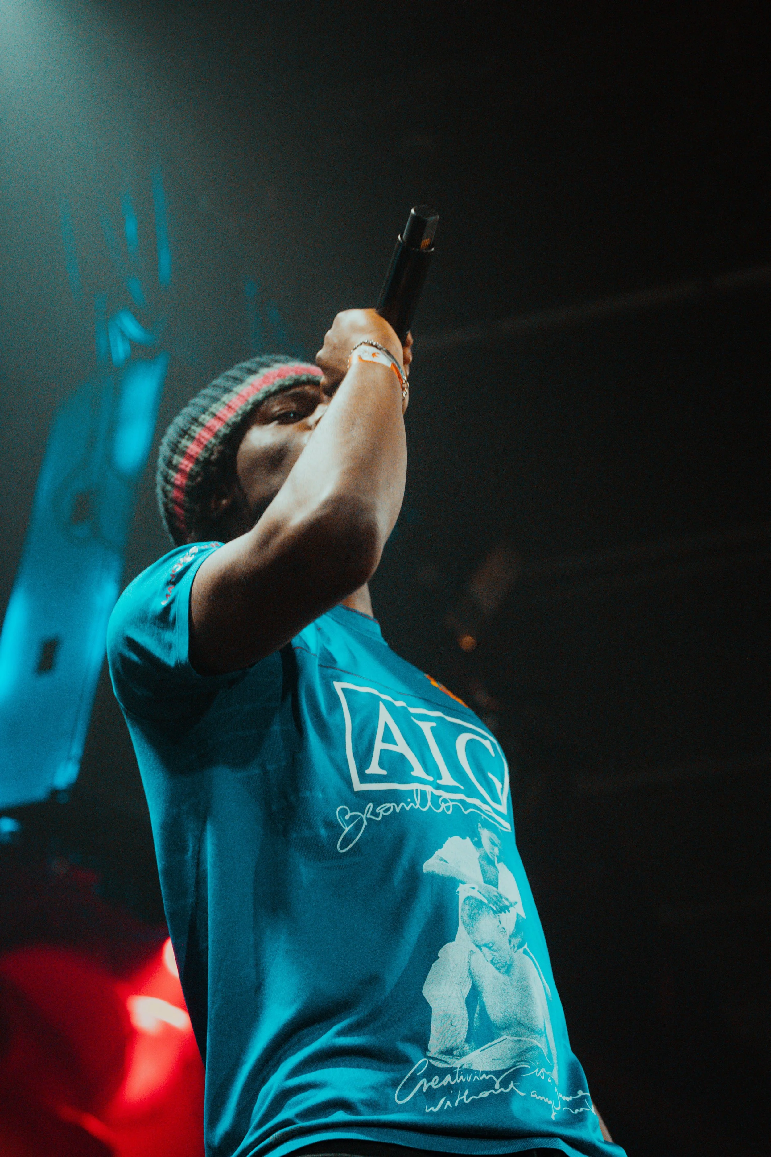 Une personne portant un t-shirt bleu et un bonnet rayé, tenant un microphone et se produisant sur scène dans des conditions de faible éclairage.