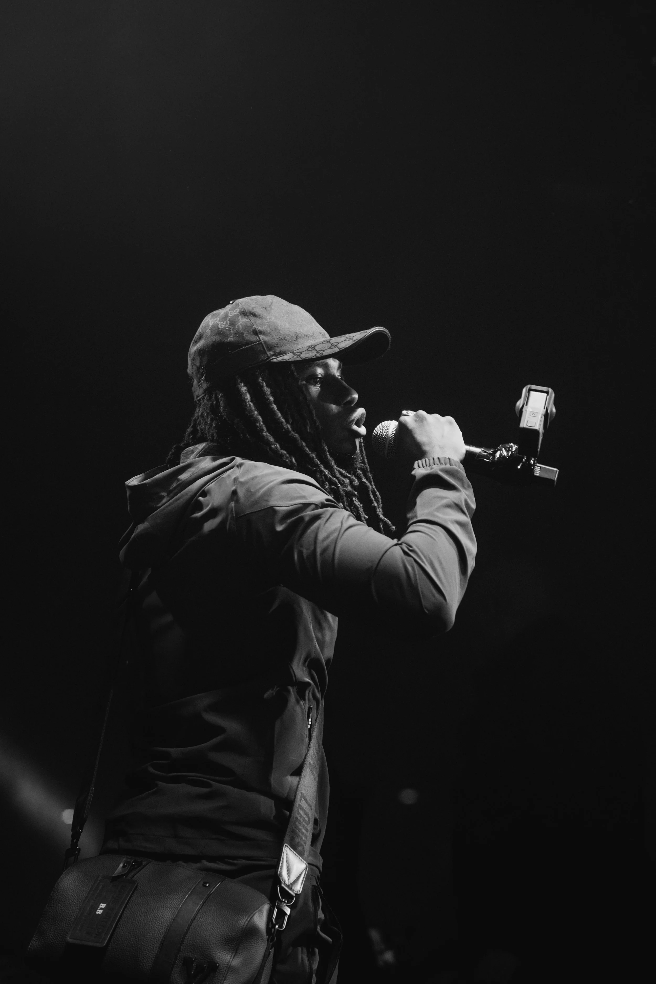 Une femme avec des dreadlocks portant une casquette et un manteau, parlant ou chantant dans un microphone dans une scène sombre.