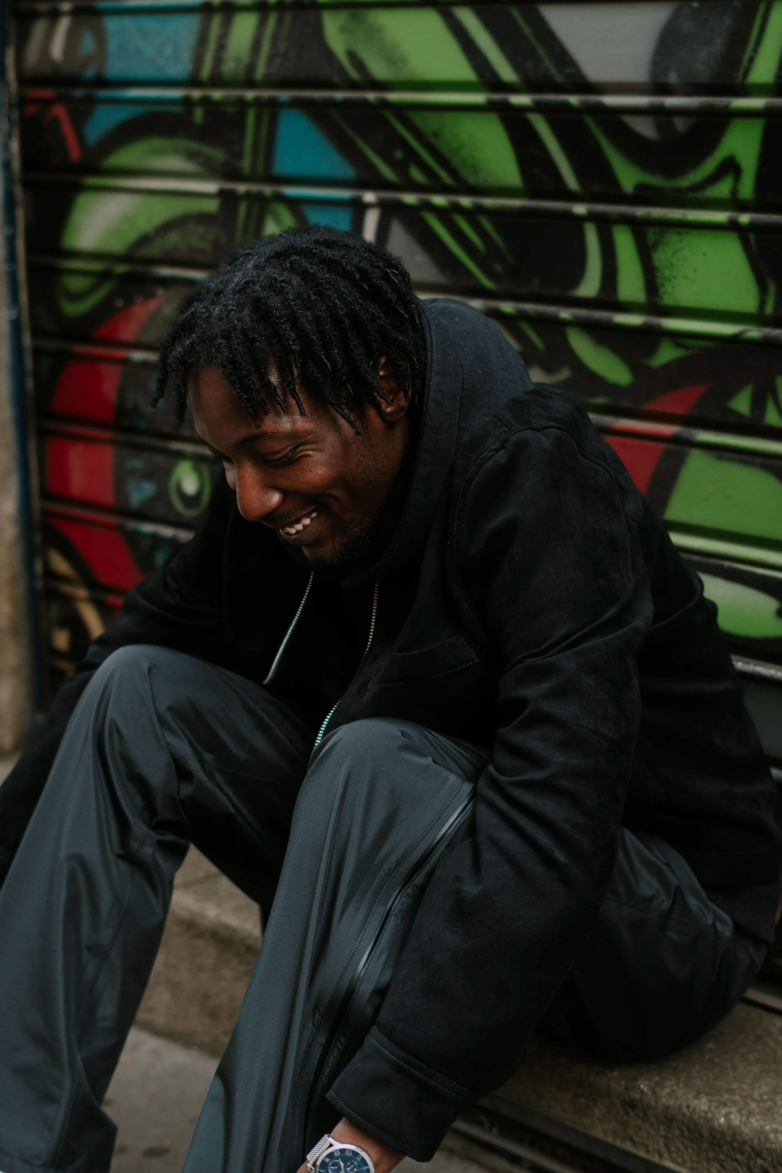 Un homme souriant, assis et en train de mettre son sac à dos, avec un fond de graffitis colorés.