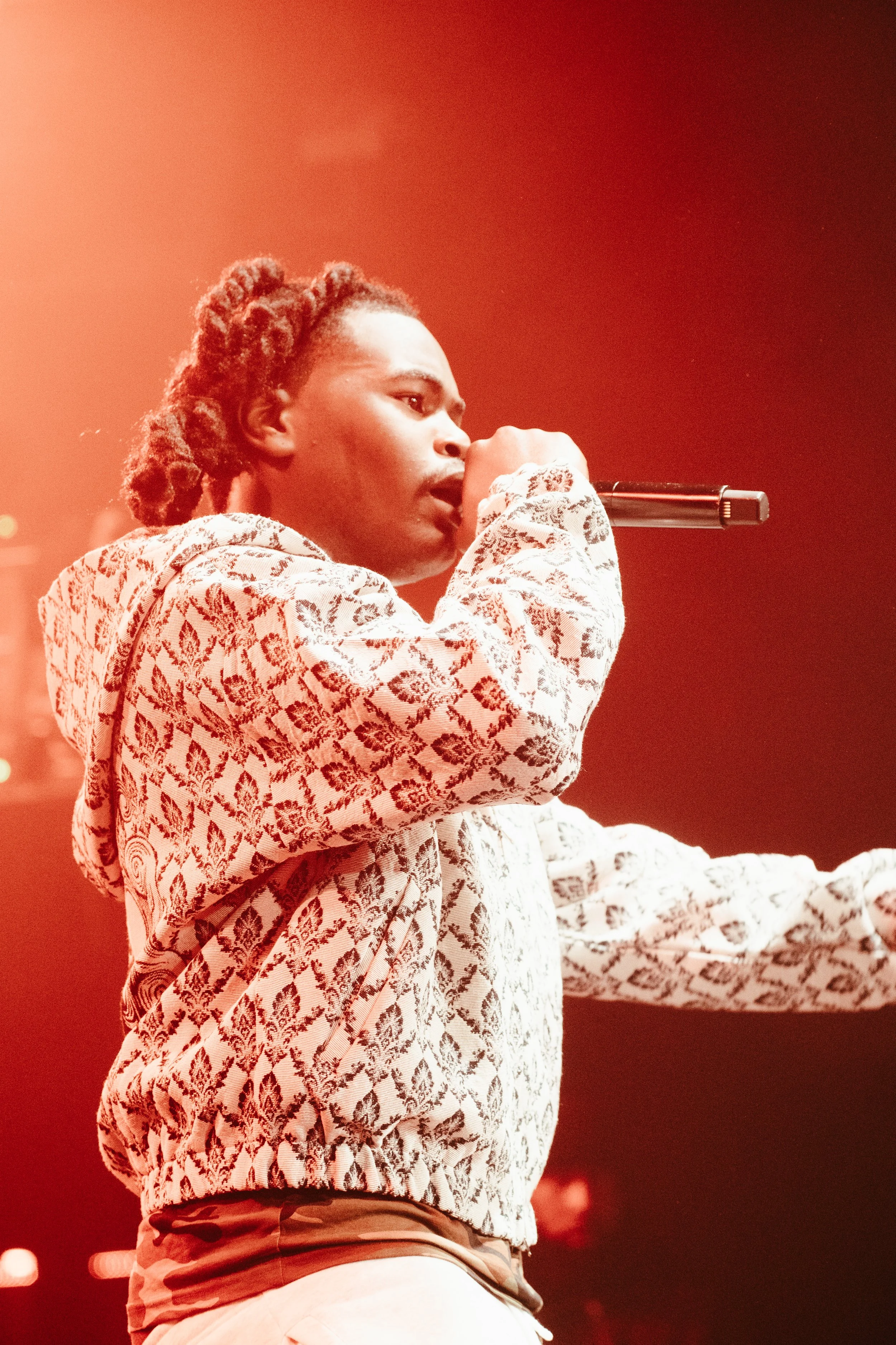 Un homme chante dans un micro lors d'un concert, portant un sweat à motif floral et se trouve sous un éclairage rouge.