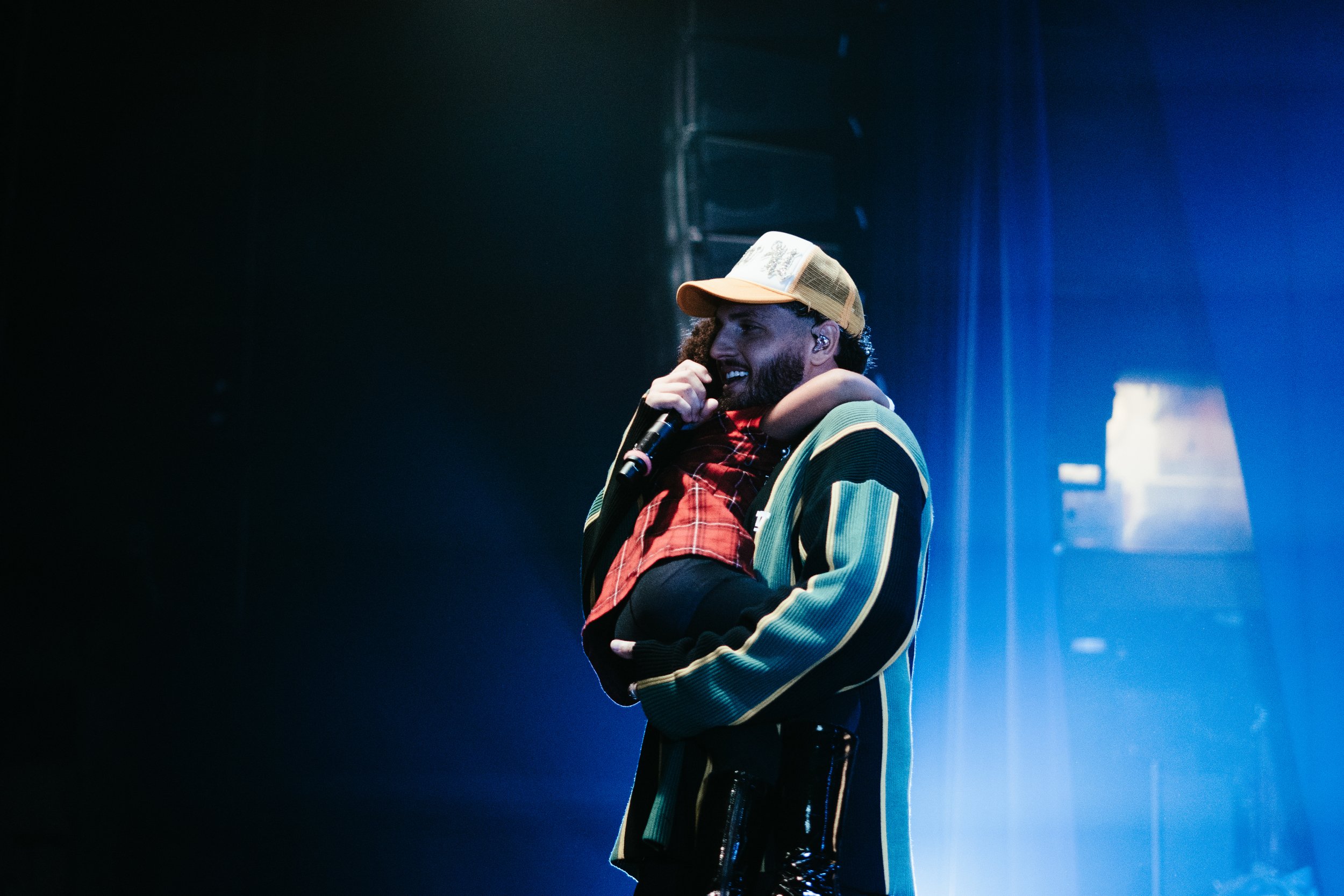 Un homme avec une barbe en costume coloré tient une jeune fille dans ses bras sur scène, tous deux souriants. L'homme porte une casquette beige, et la scène est éclairée en bleu.