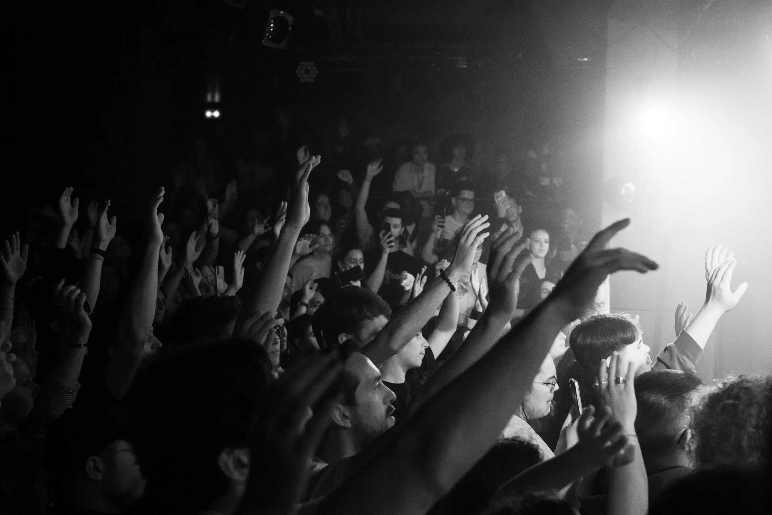 Un public dans une salle de concert ou un événement, levant la main, avec un éclairage intense venant d'une scène ou d'une source lumineuse à droite.