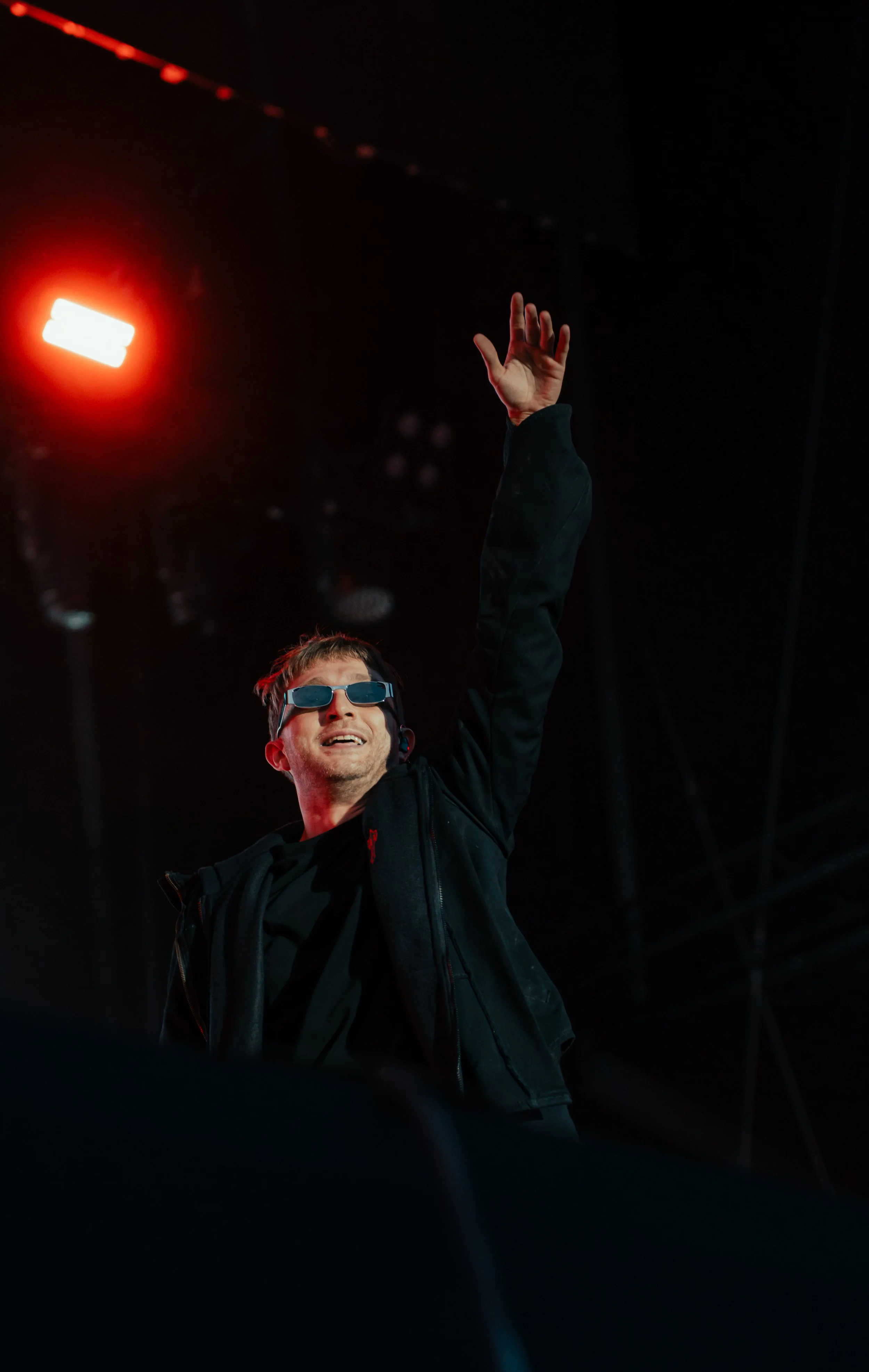 Un homme portant des lunettes de soleil noires, un vêtement noir et un casque parlant, souriant et levant la main lors d'une performance ou d'un événement en intérieur.
