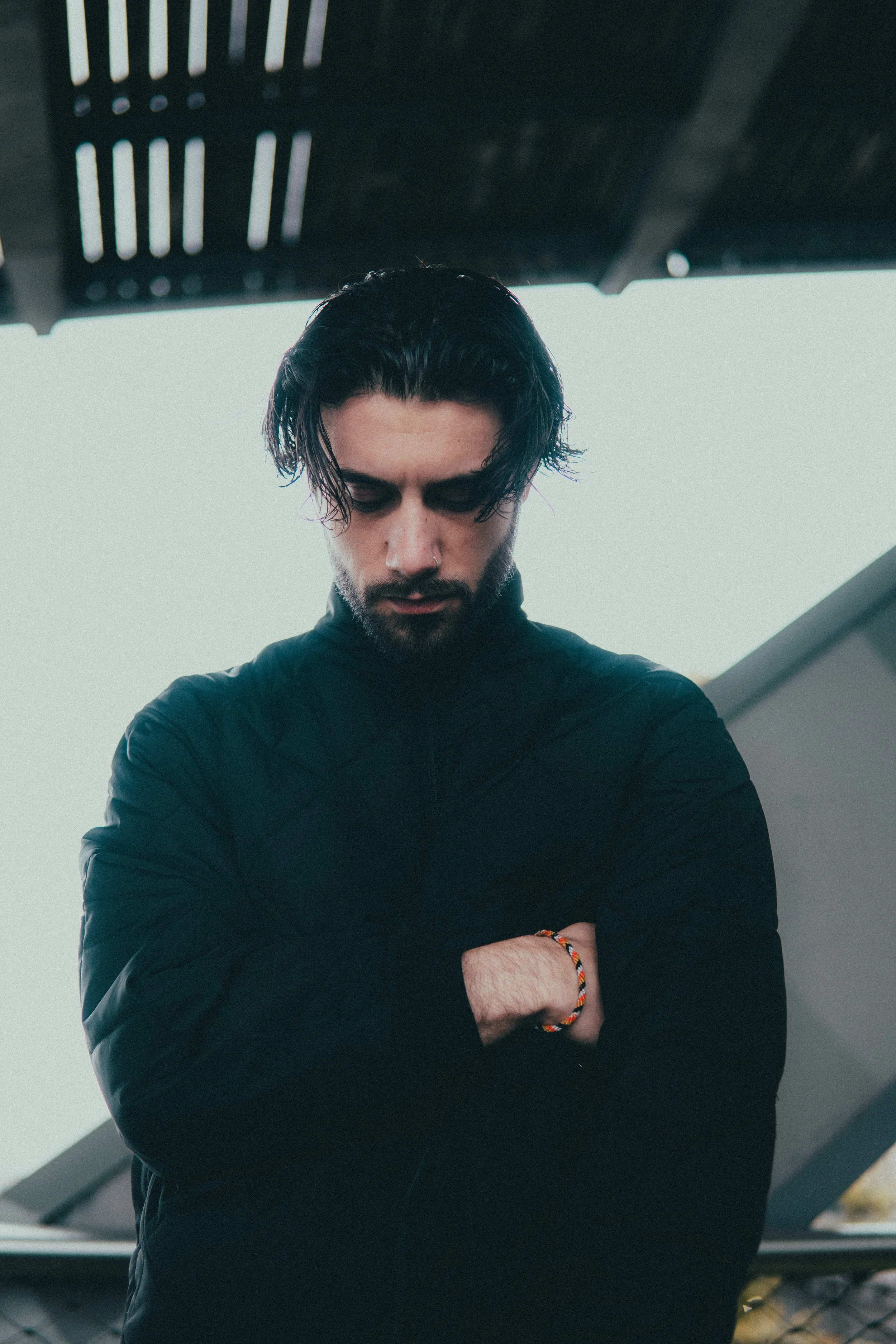 Jeune homme aux cheveux longs et coiffé en arrière, portant une veste noire, croisant les bras, sous un toit en bois avec un fond clair.