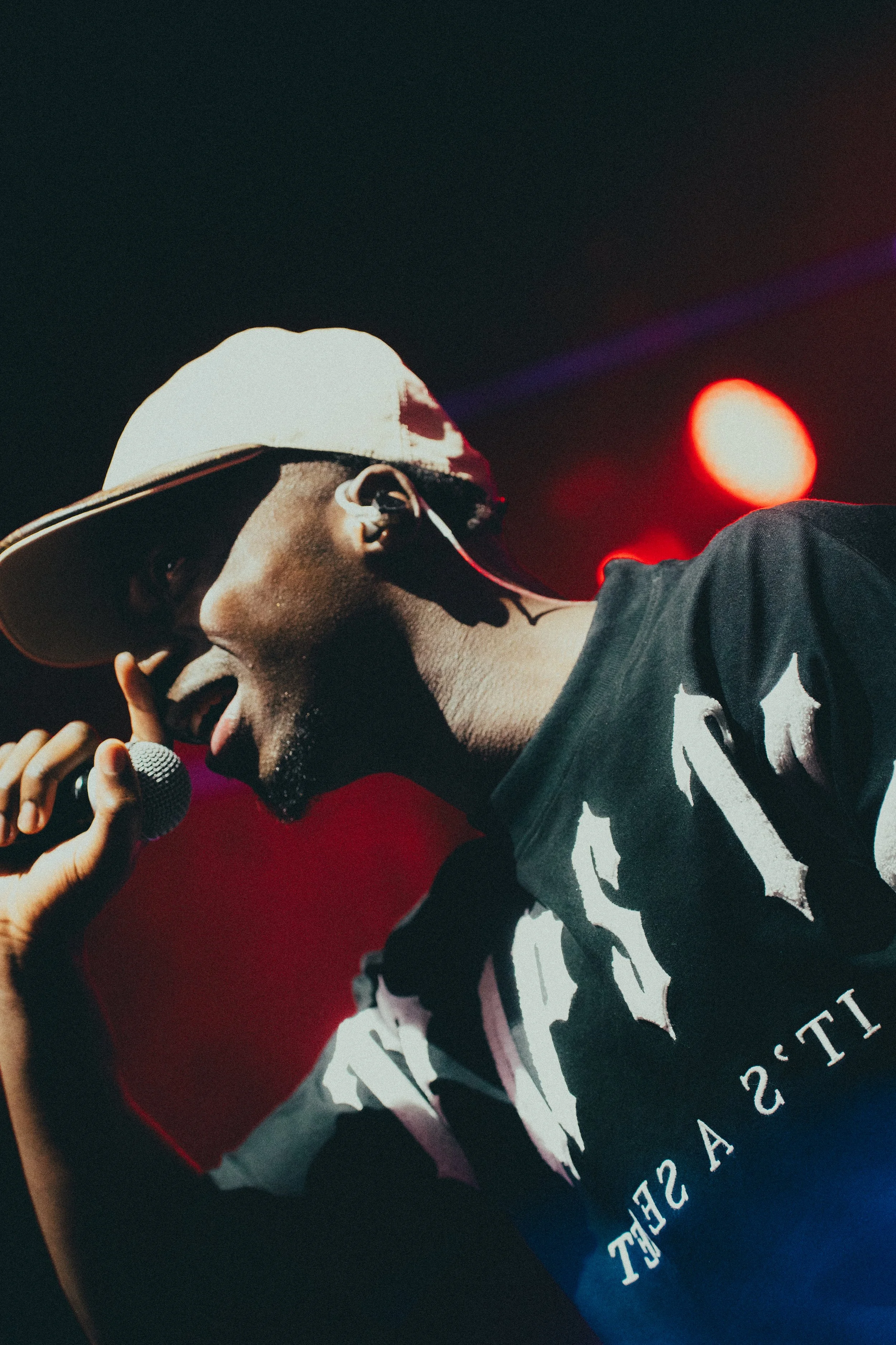 Un homme porte un bonnet blanc, parle ou chante dans un micro, avec un arrière-plan de lumière rouge et un fond sombre.