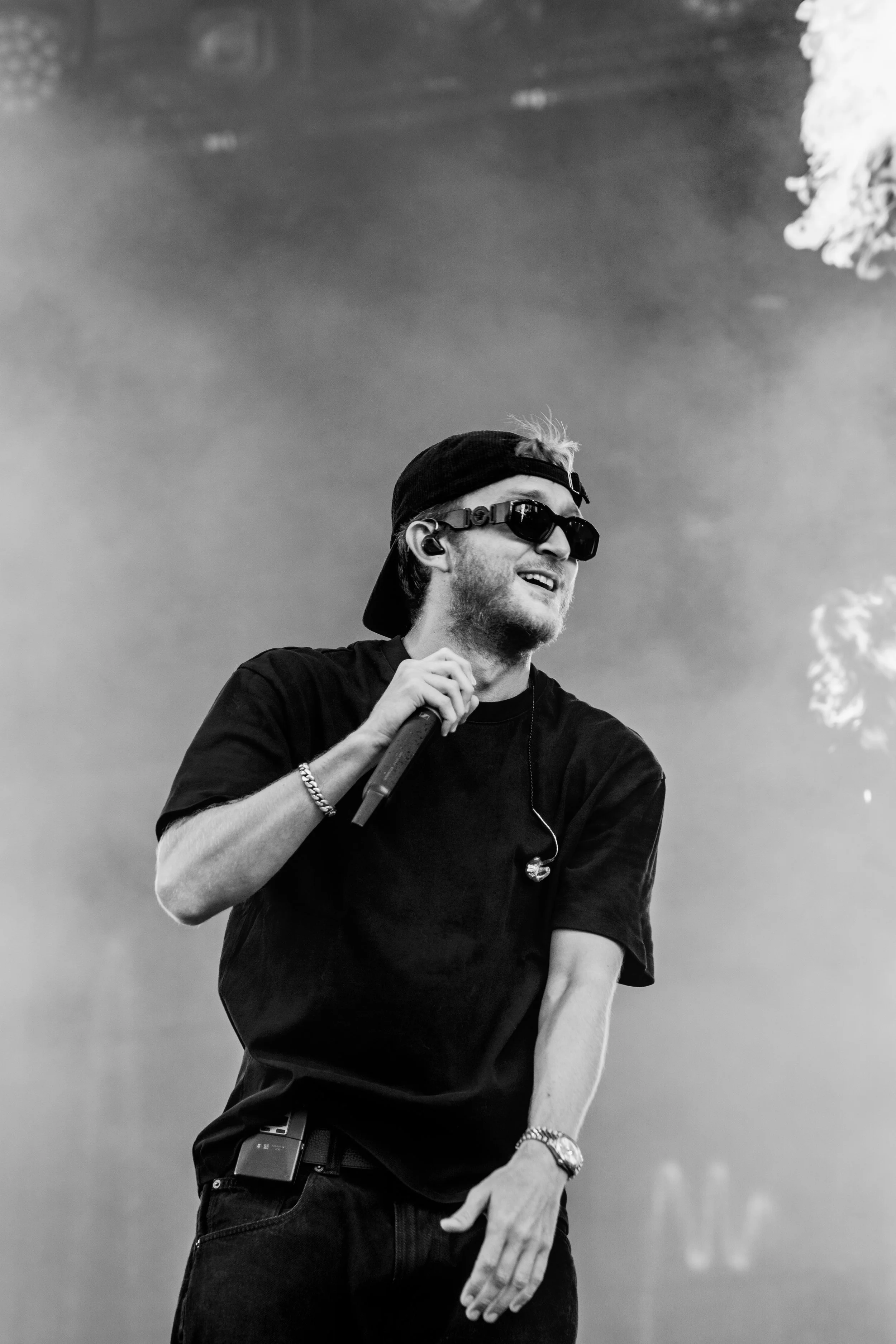 Un homme portant une casquette, des lunettes de soleil et une montre, tenant un microphone, semble être en train de performer sur scène.