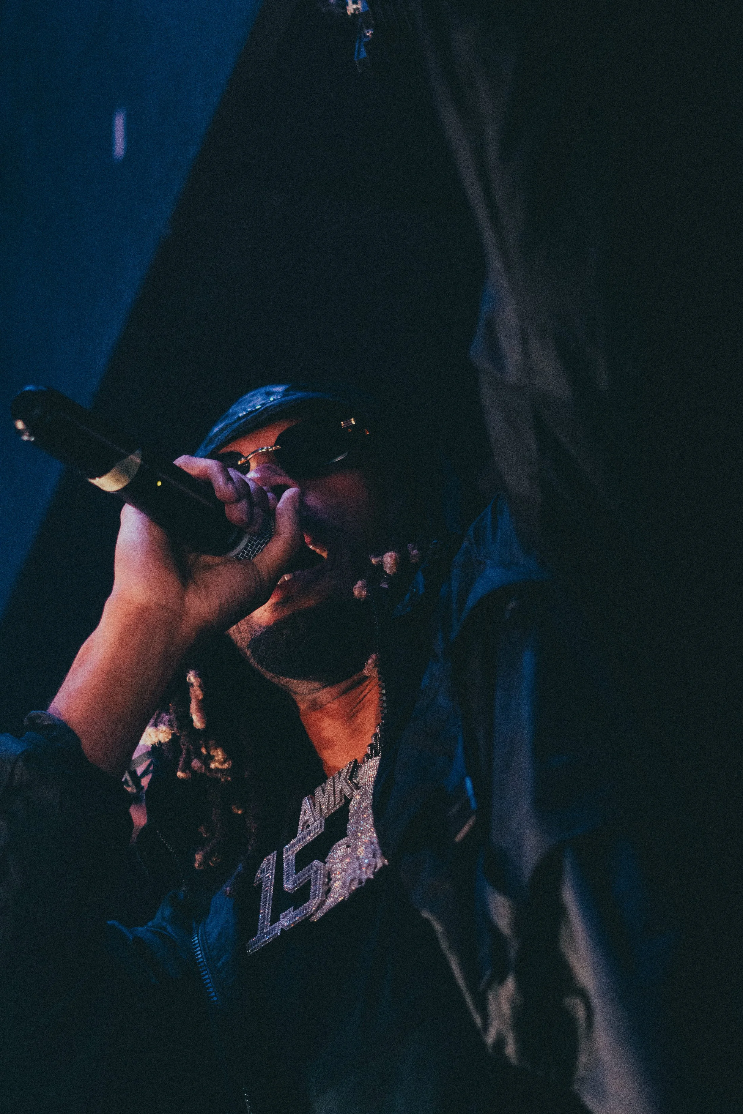 Un artiste de rap en train de chanter dans un micro lors d'une performance en concert, portant des lunettes de soleil, une veste noire et un bijoux avec le chiffre 15.