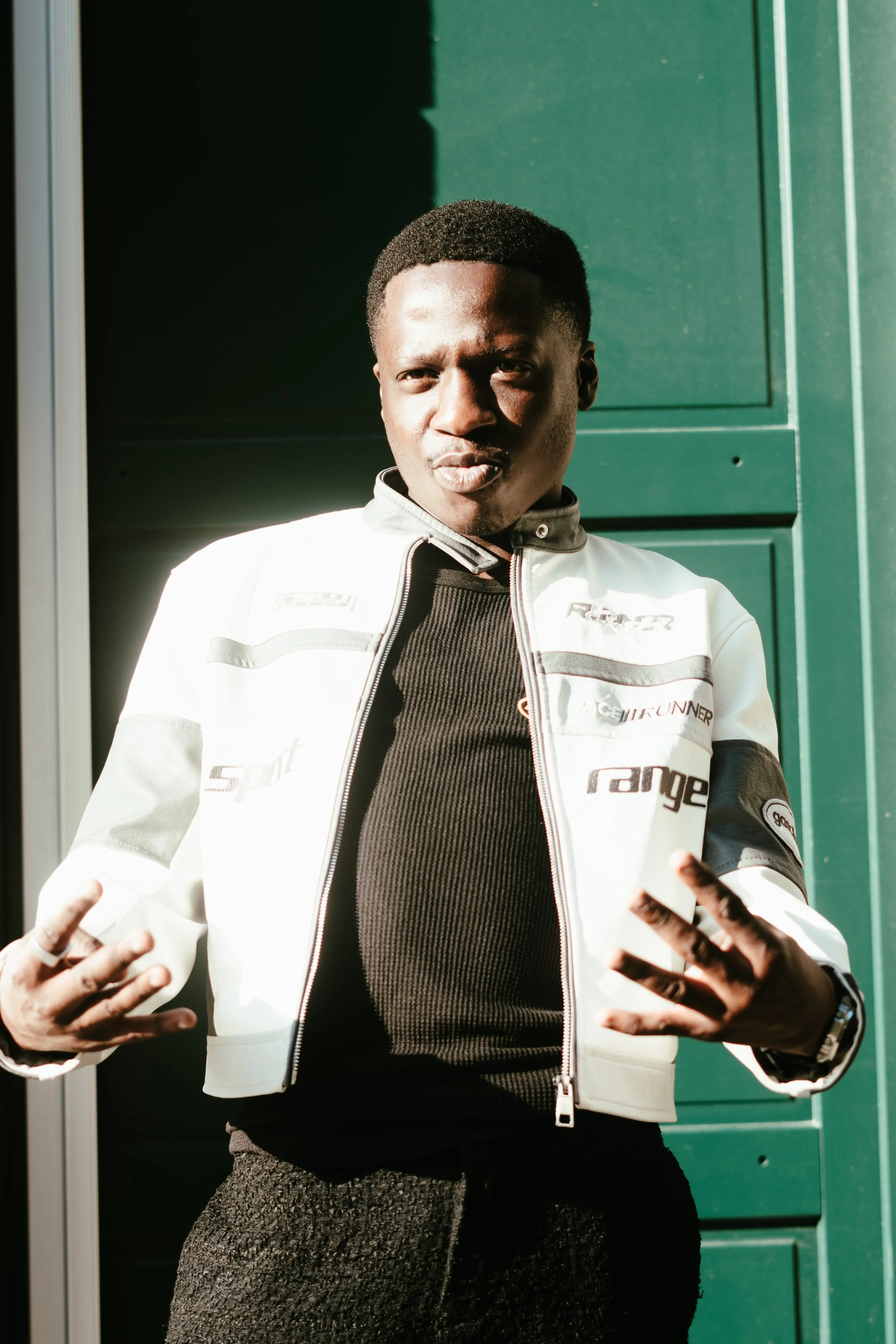 Un jeune homme en veste de moto blanche pose devant un mur vert, faisant un geste avec ses mains.