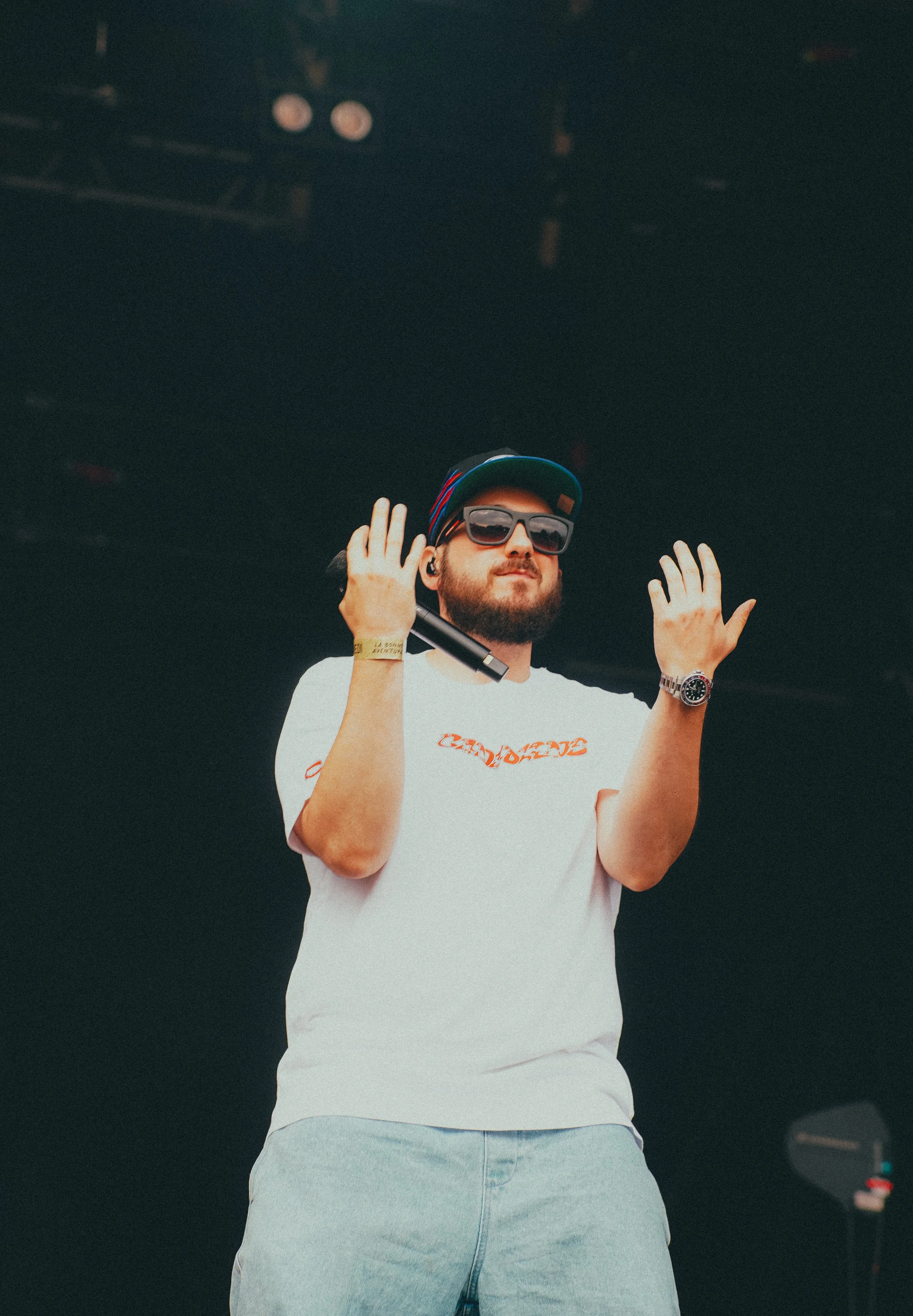 Un homme portant des lunettes de soleil, une casquette et un t-shirt blanc se tenant sur scène, avec une main en train de faire un geste dans l'air.