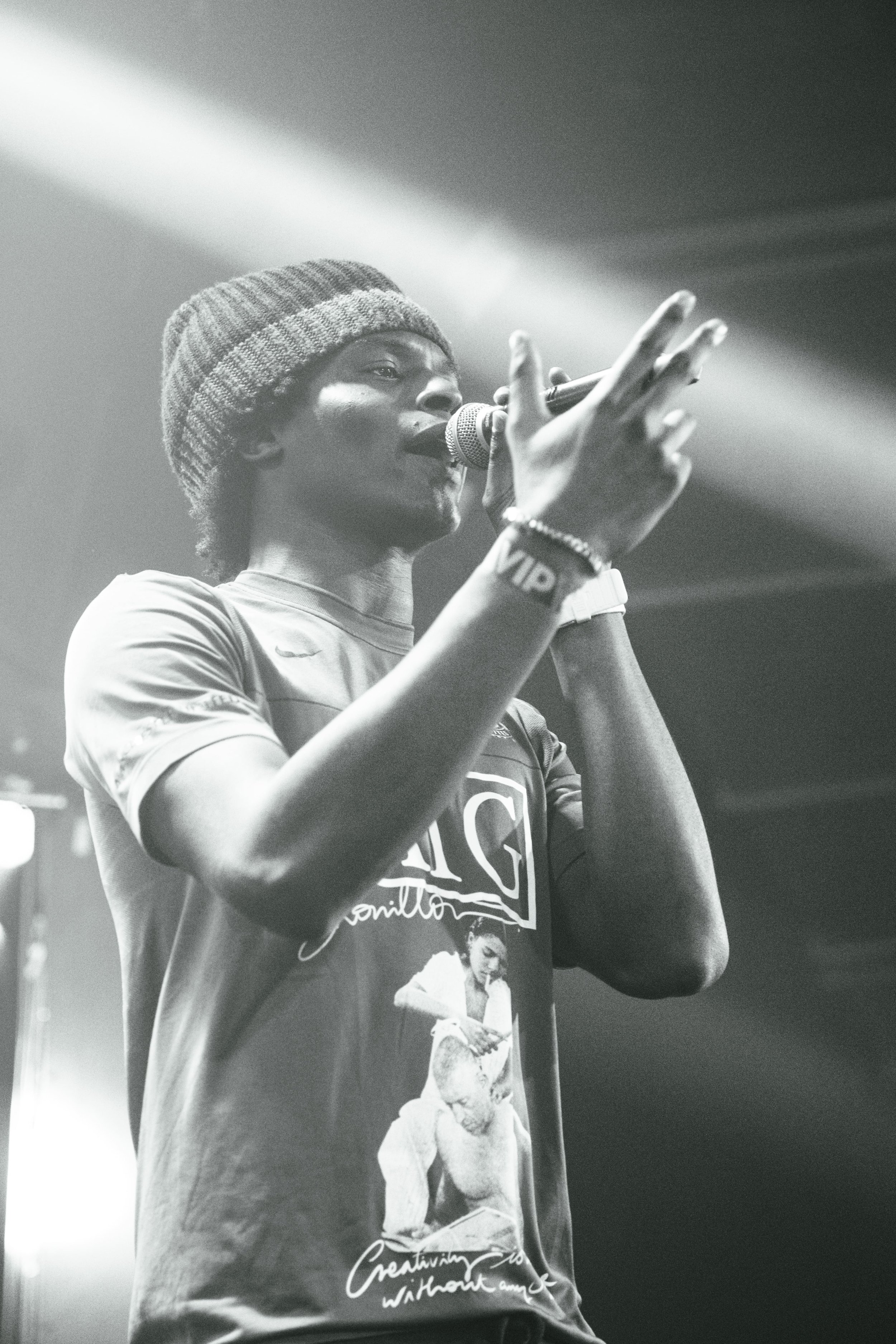 Une artiste noire chantant dans un micro, portant un bonnet en laine, un bracelet, une montre, un t-shirt avec une image et des inscriptions, sur scène avec un éclairage en arrière-plan.