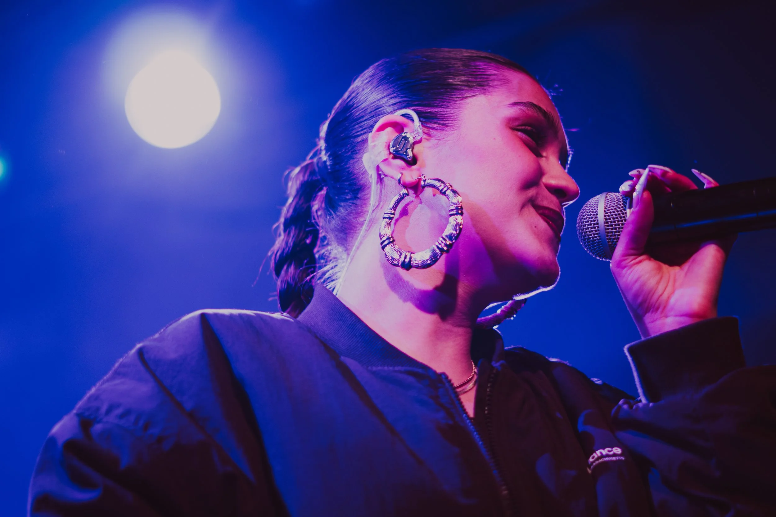 Femme chantant ou parlant dans un micro lors d'une performance, avec des lumières colorées en arrière-plan.