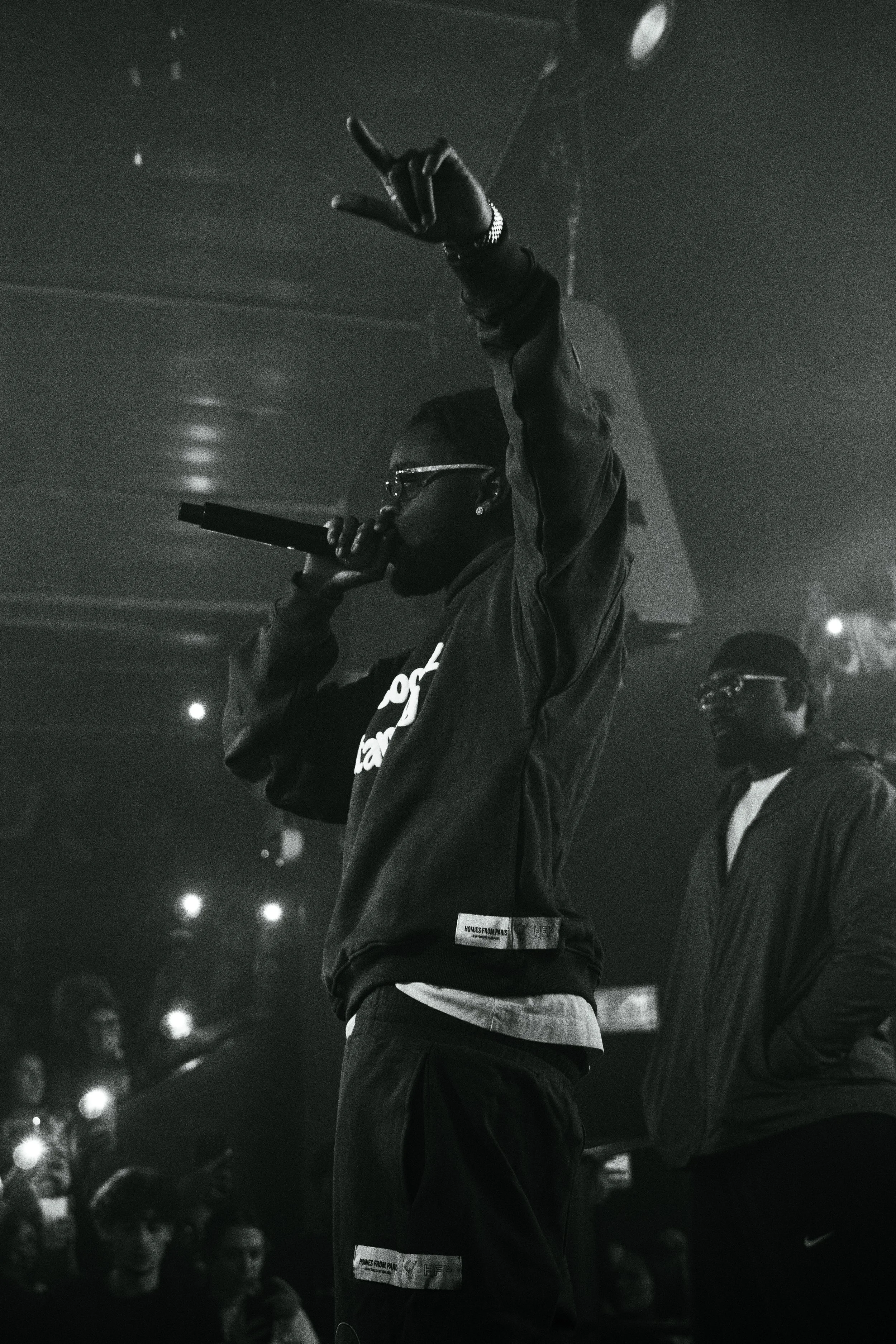 Un artiste rappeur porte des lunettes en train de performer sur scène, tenant un micro dans une main et levant l'autre main, lors d'un concert ou d'une spectacle.