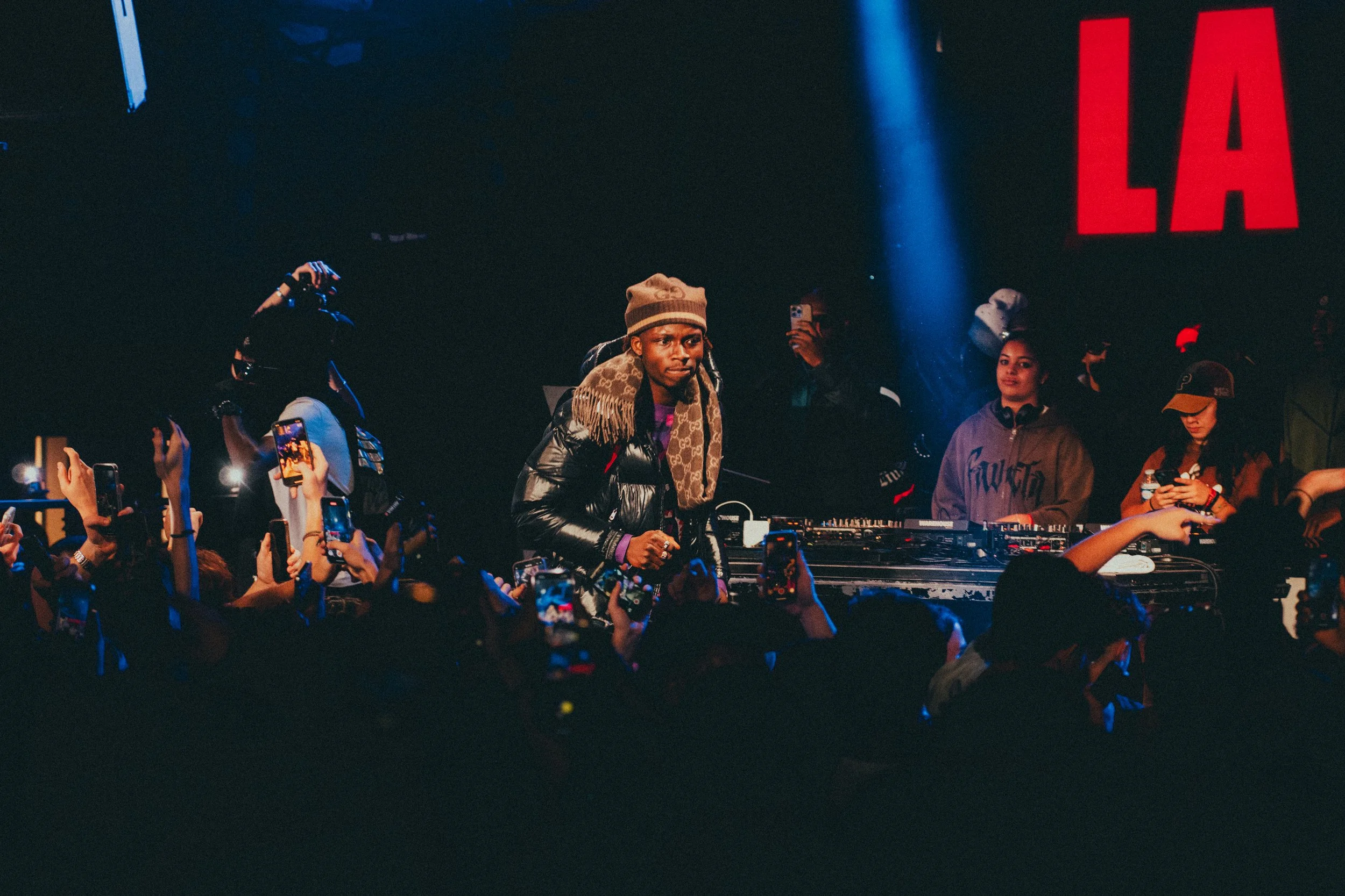 DJ en train de mixer lors d'un concert ou d'une fête, entouré de spectateurs prenant des photos, avec un grand panneau rouge où il est écrit 'LA' en arrière-plan.