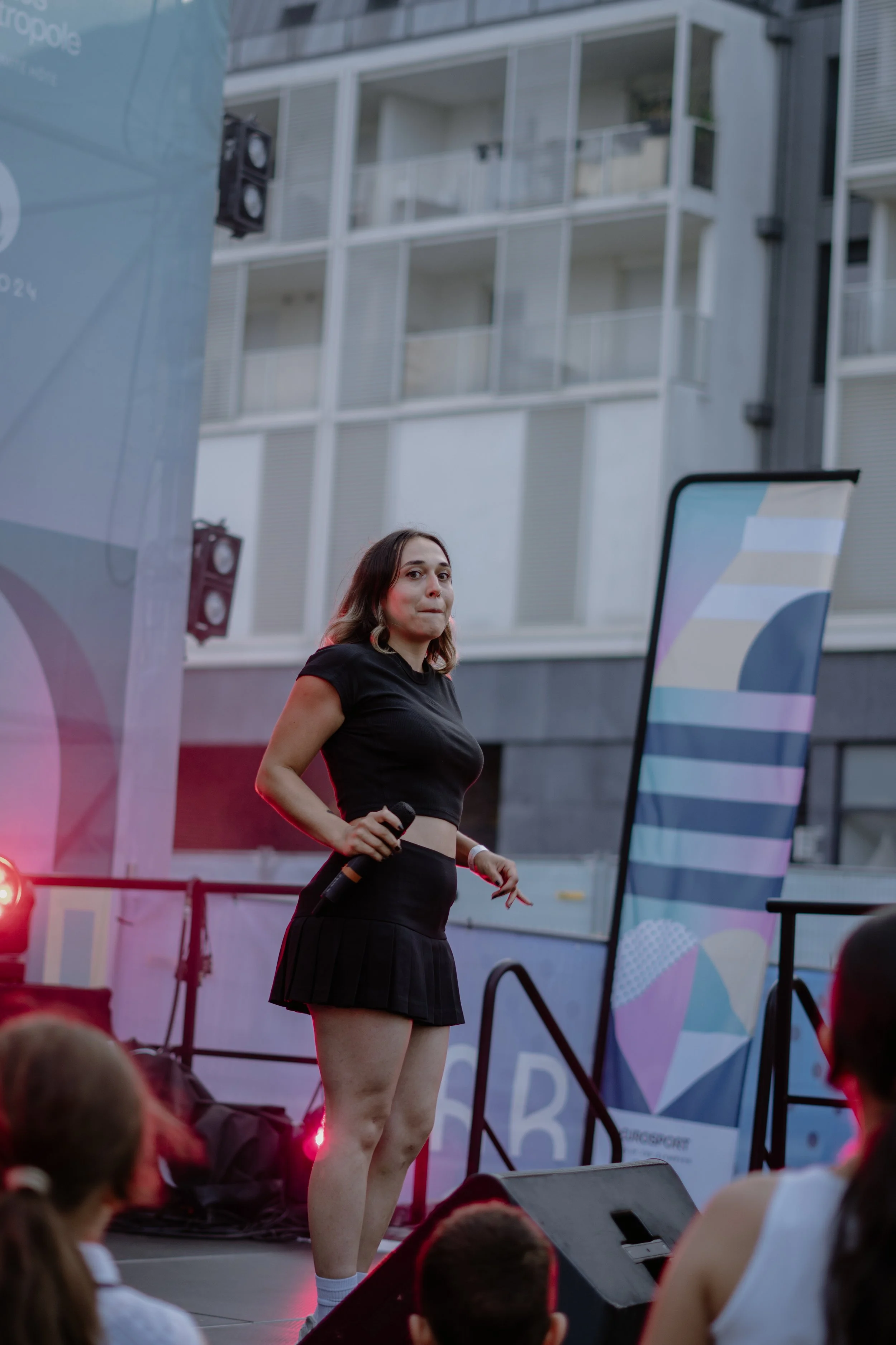 Une femme chante avec un micro sur scène lors d'un événement en extérieur, avec un bâtiment moderne en arrière-plan.