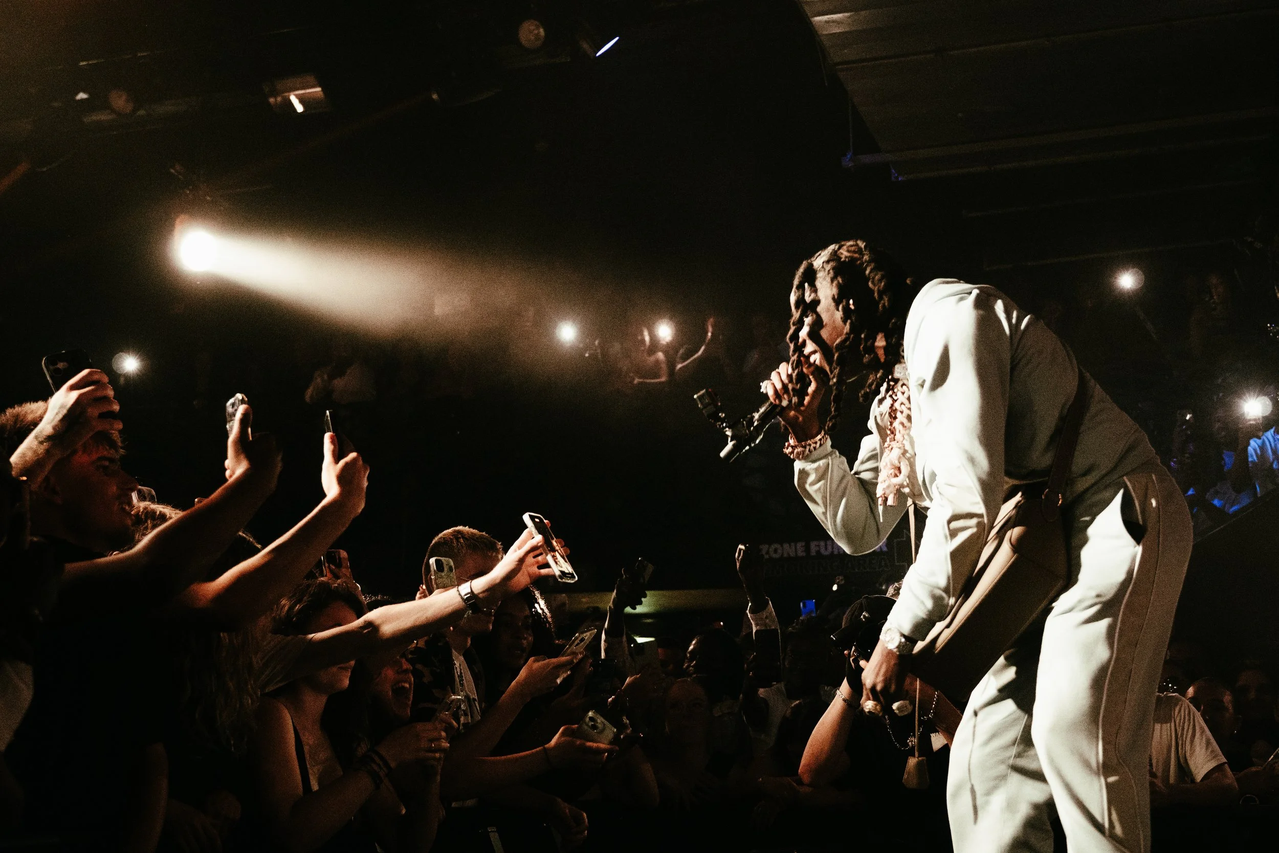 Un artiste avec des dreadlocks chante dans un micro sur scène lors d'un concert, face à un public enthousiaste qui prend des photos avec leurs téléphones, dans un lieu sombre avec des lumières dirigées vers la scène.