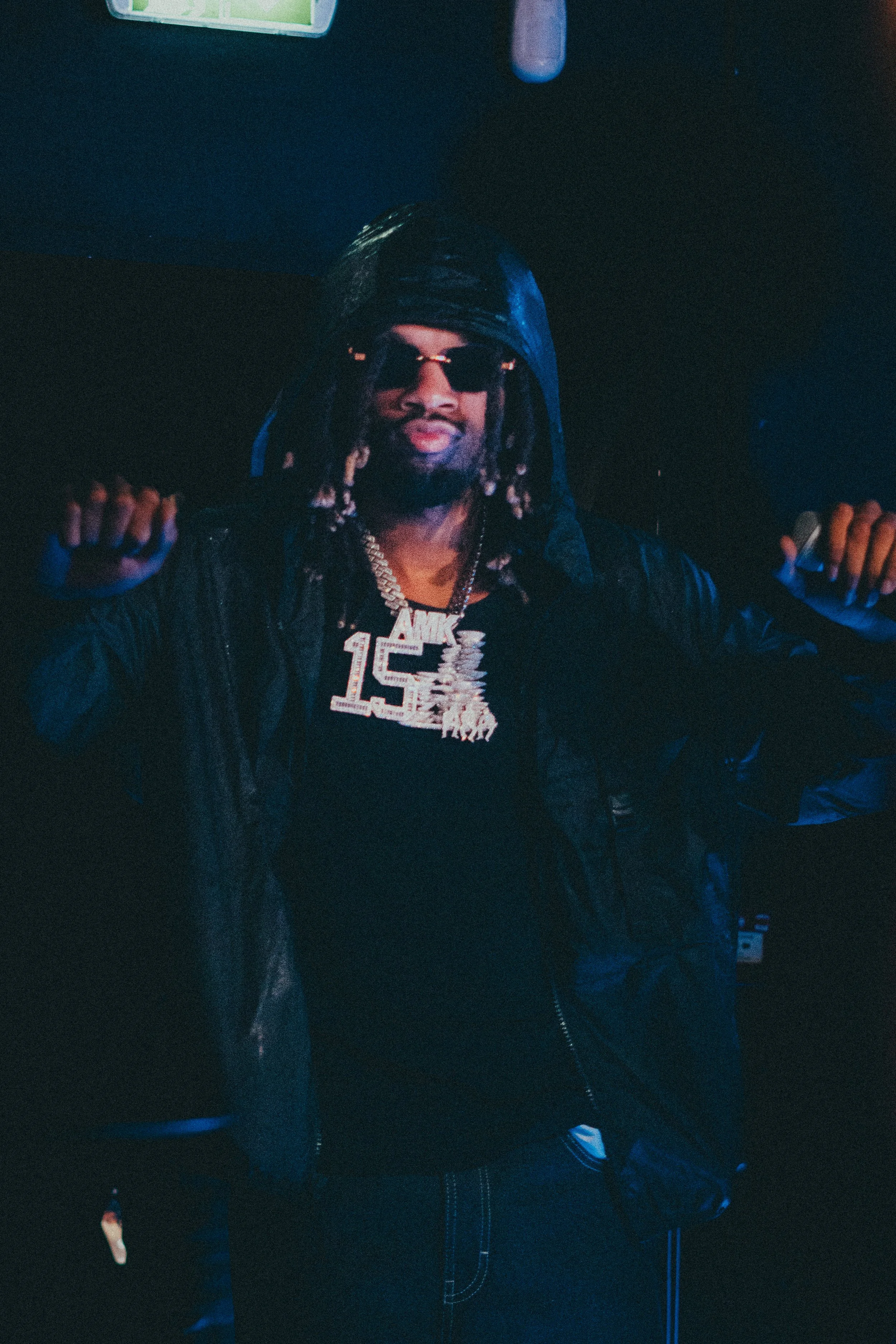 Un homme portant des lunettes de soleil, un sweat à capuche et un gros collier en or avec la mention "15" posant dans un environnement sombre.