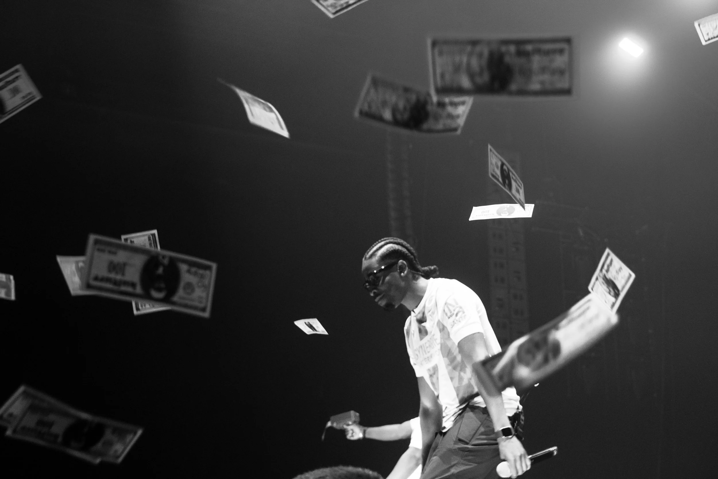 Une personne portant des lunettes de soleil, un t-shirt blanc, et une coiffure avec des tresses, se tient parmi des billets de banque en vol dans un environnement sombre.