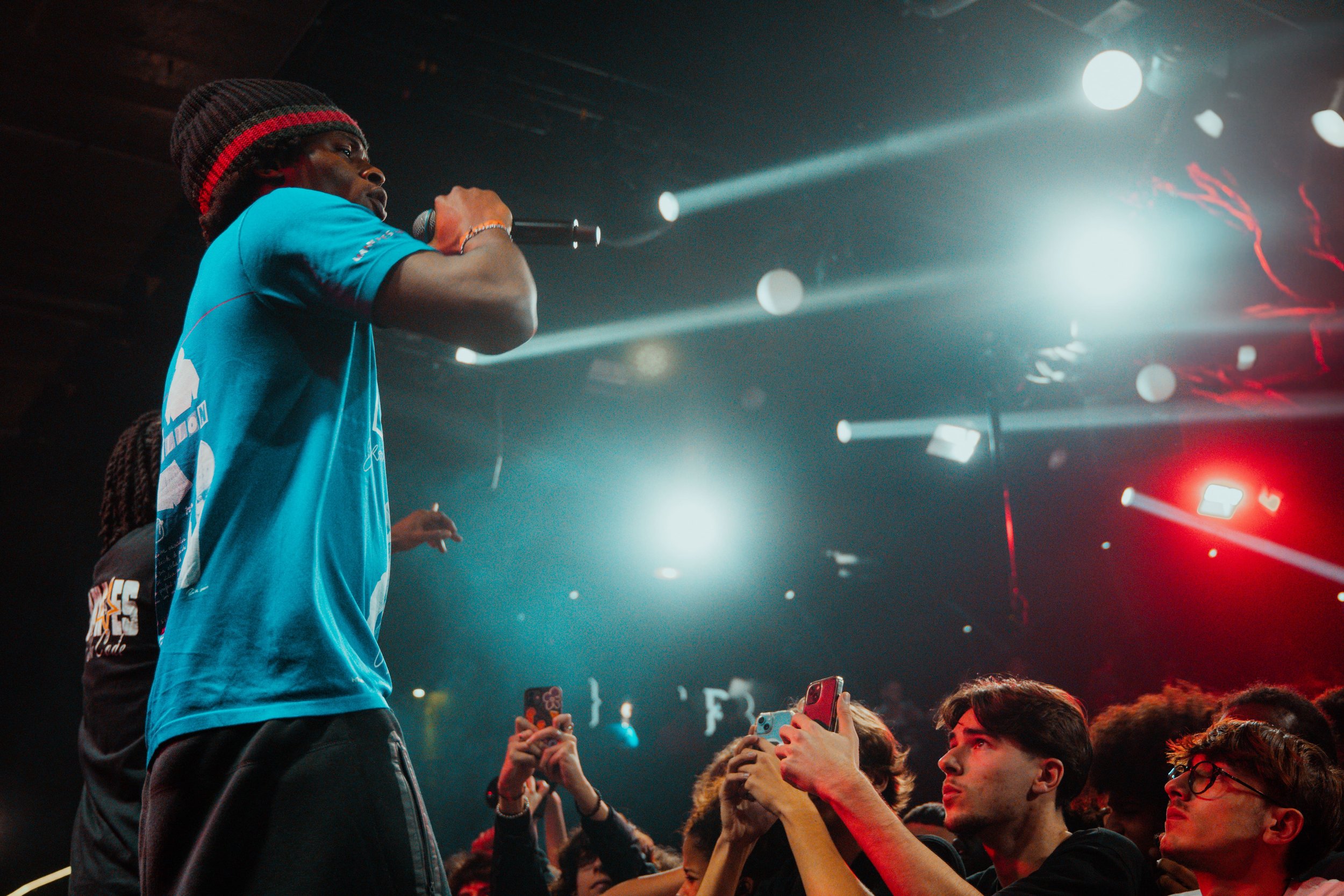 Un artiste avec un chapeau en train de chanter lors d'un spectacle devant un public qui enregistre avec des téléphones.