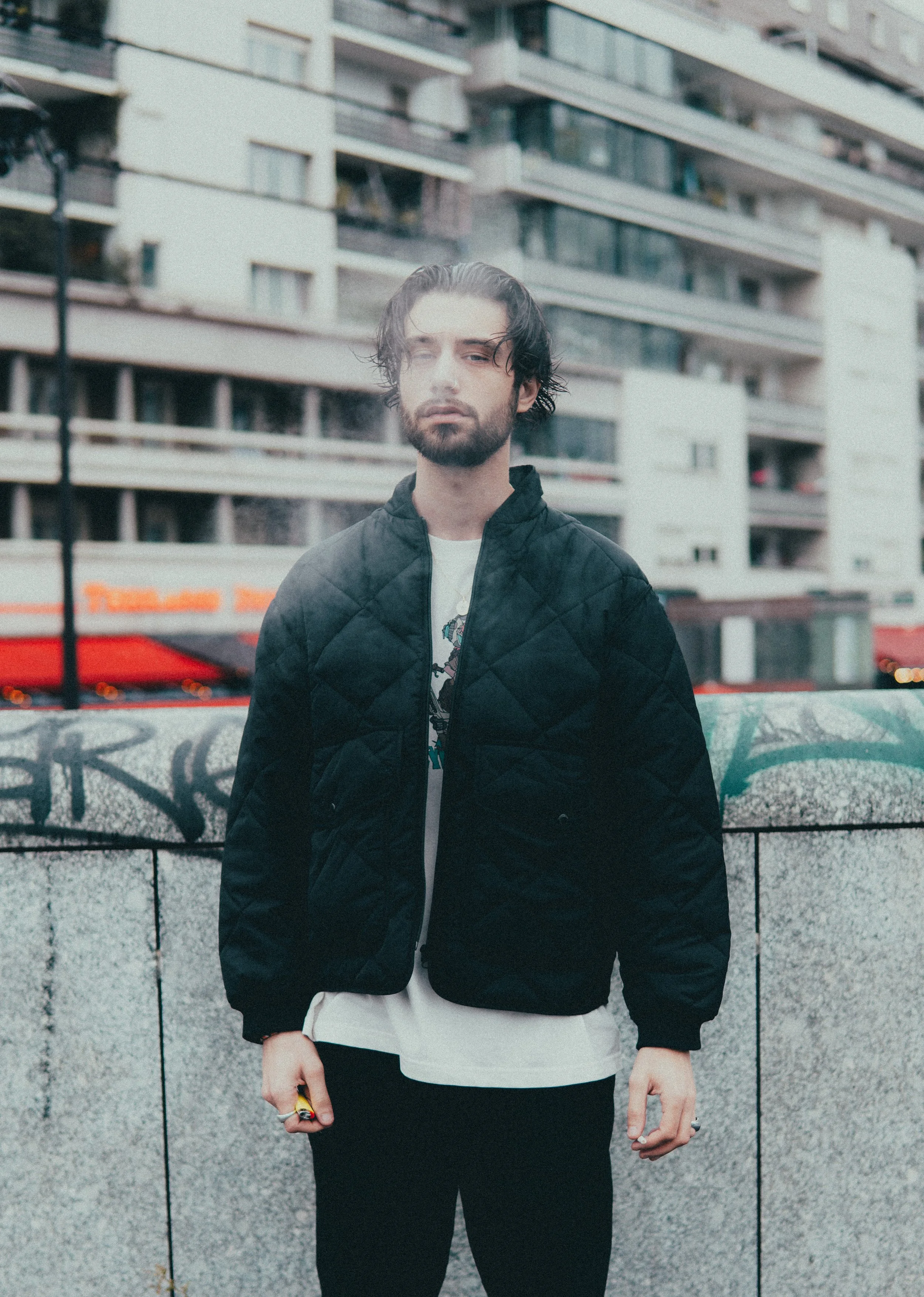 Un jeune homme avec des cheveux bruns et une barbe porte une veste noire en ville, devant un mur graffiti et un immeuble résidentiel