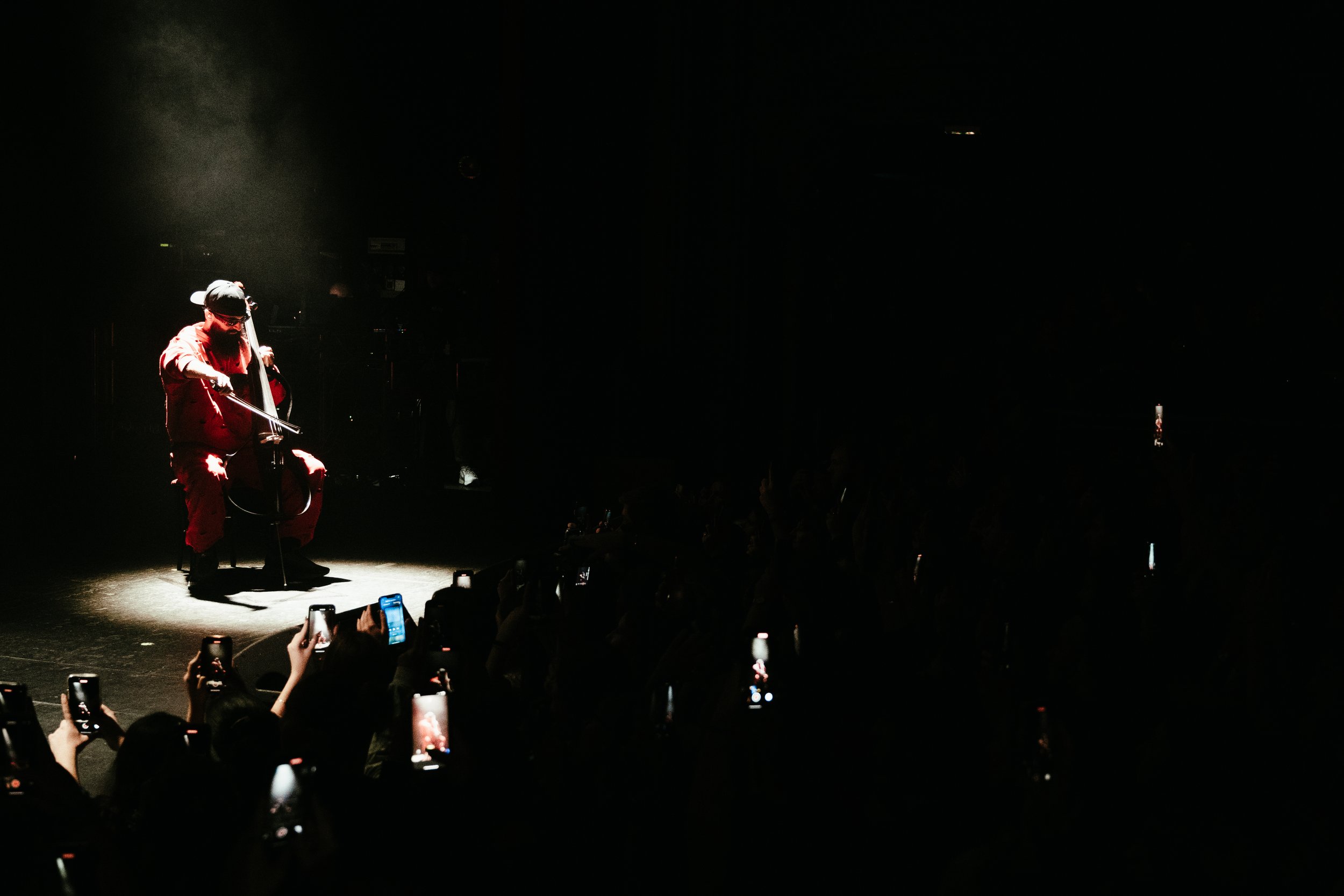 Un musicien joue du violoncelle sur scène, éclairé par un faisceau lumineux, devant un public nombreux qui prend des photos avec leurs téléphones.