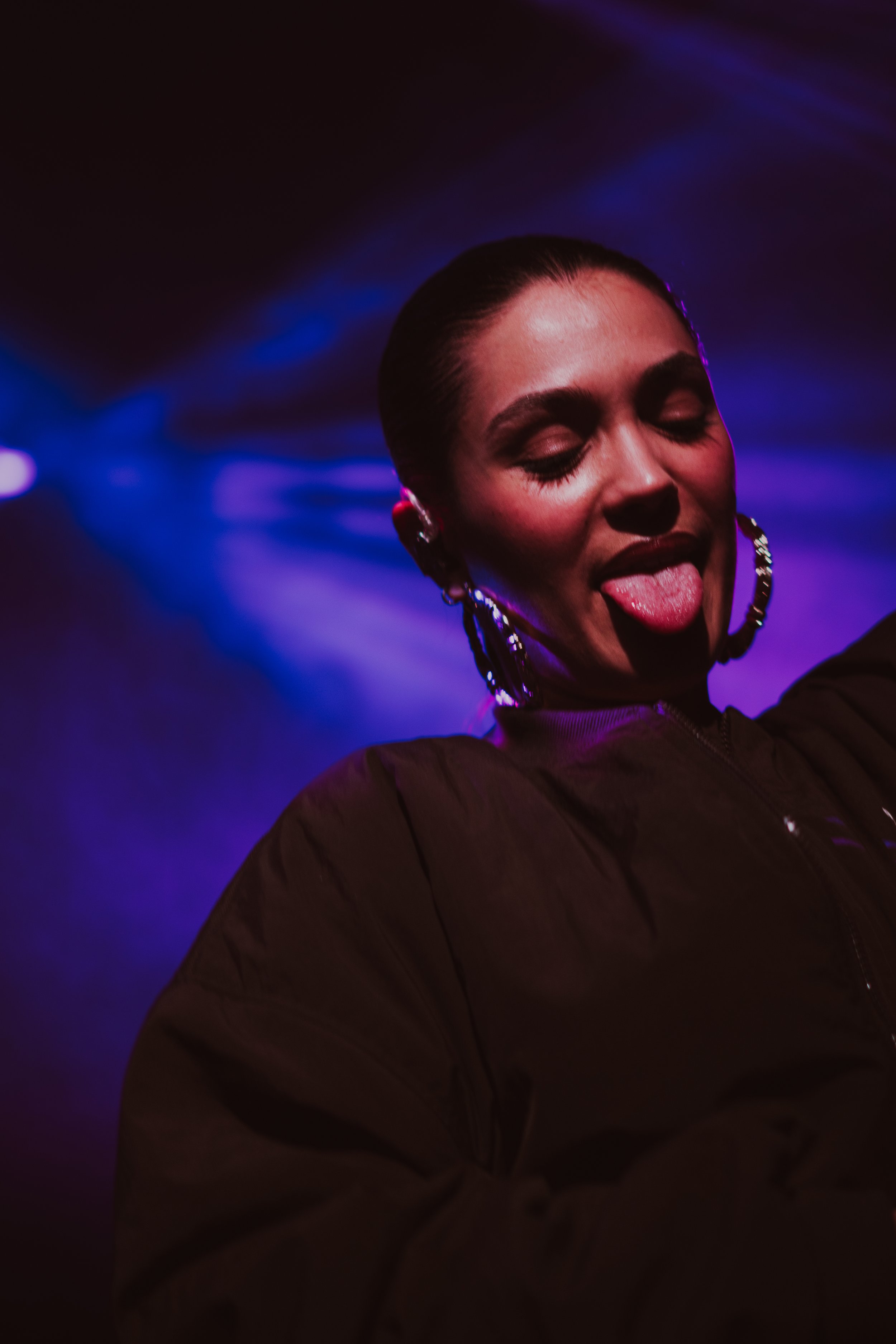 Une femme avec des boucles d'oreilles et un maquillage sombre, fait un geste avec la langue dans un environnement sombre avec des lumières violettes et bleues.