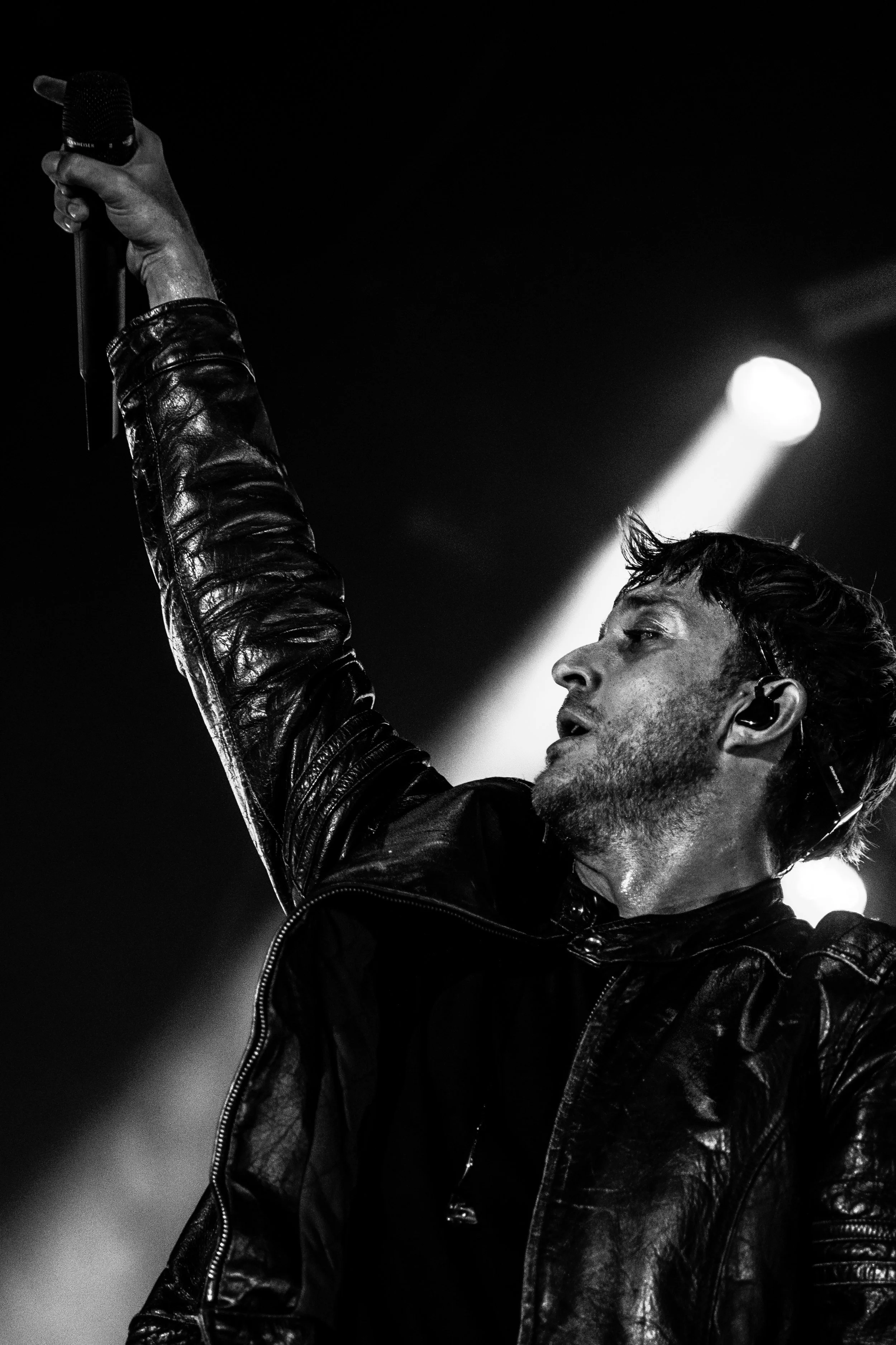 Un homme portant une veste en cuir noire, tenant un microphone en l'air lors d'une performance sur scène. La scène est sombre avec une lumière brillante en arrière-plan.